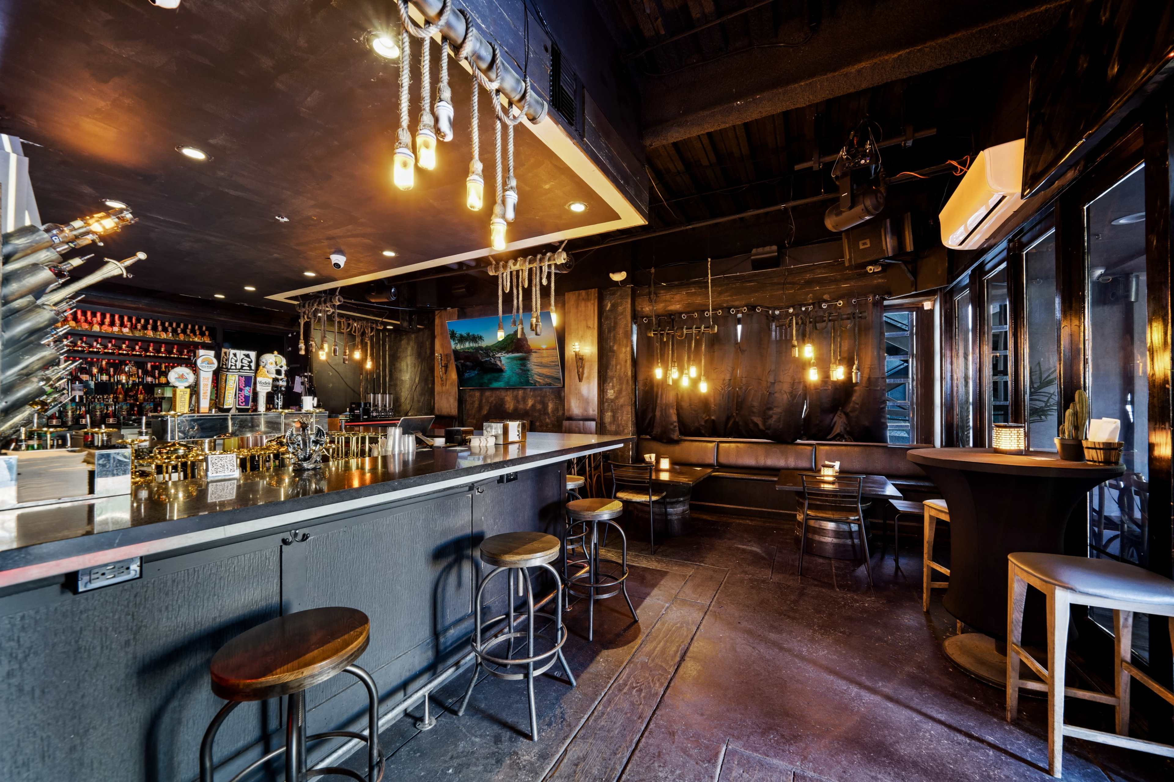 A dimly lit bar features a sleek counter with stools, a selection of bottles on shelves, and a seating area with a large window.