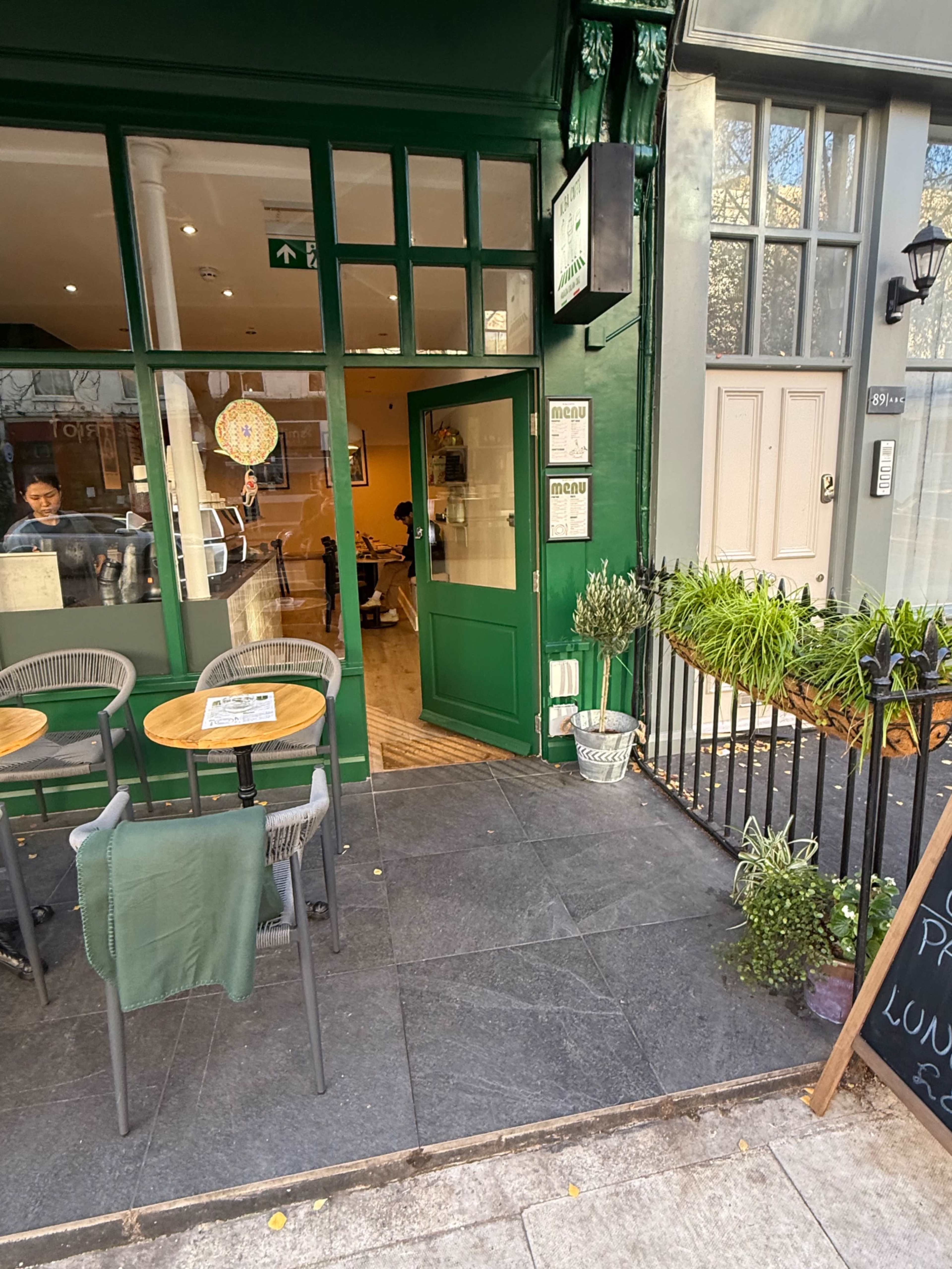A small café entrance features green walls and plants, with tables set outside and a chalkboard menu nearby.