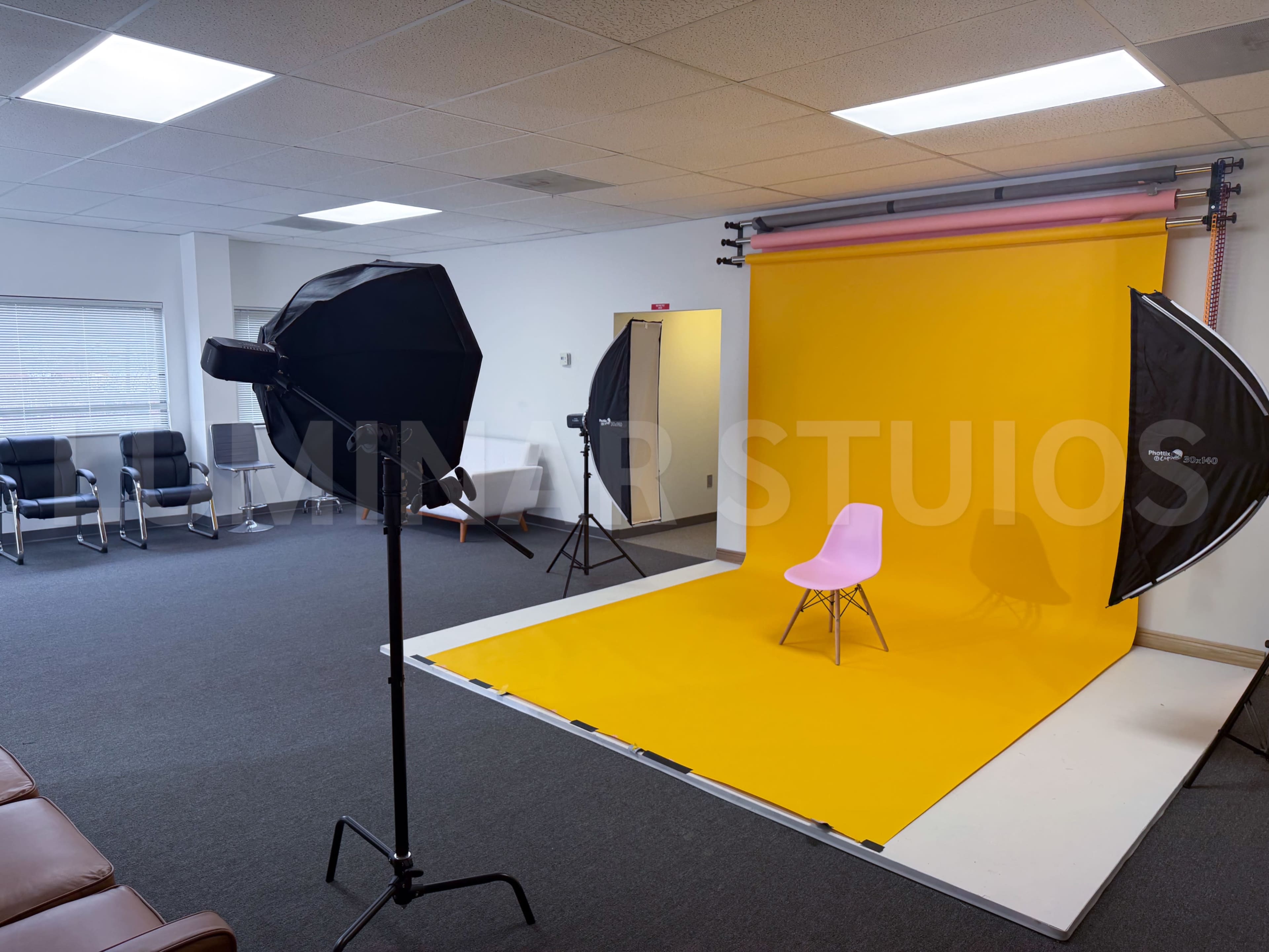 The image shows a photography studio with a yellow backdrop, a pink element above it, a pink chair in the center, and two softbox lights positioned on either side.