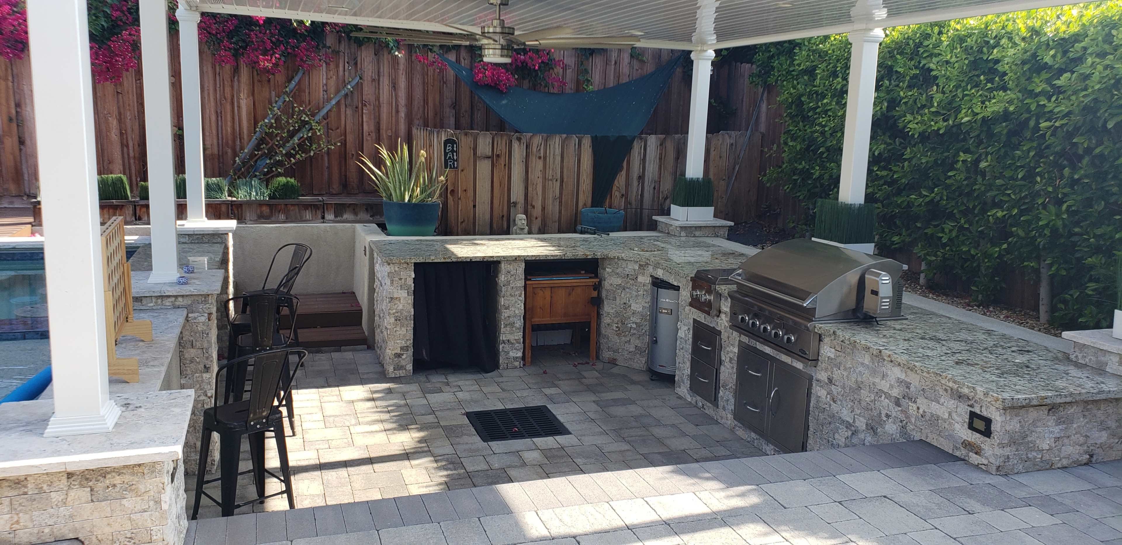 The image shows an outdoor kitchen area with a barbecue grill, countertop, and seating next to a pool, surrounded by greenery and flowers.