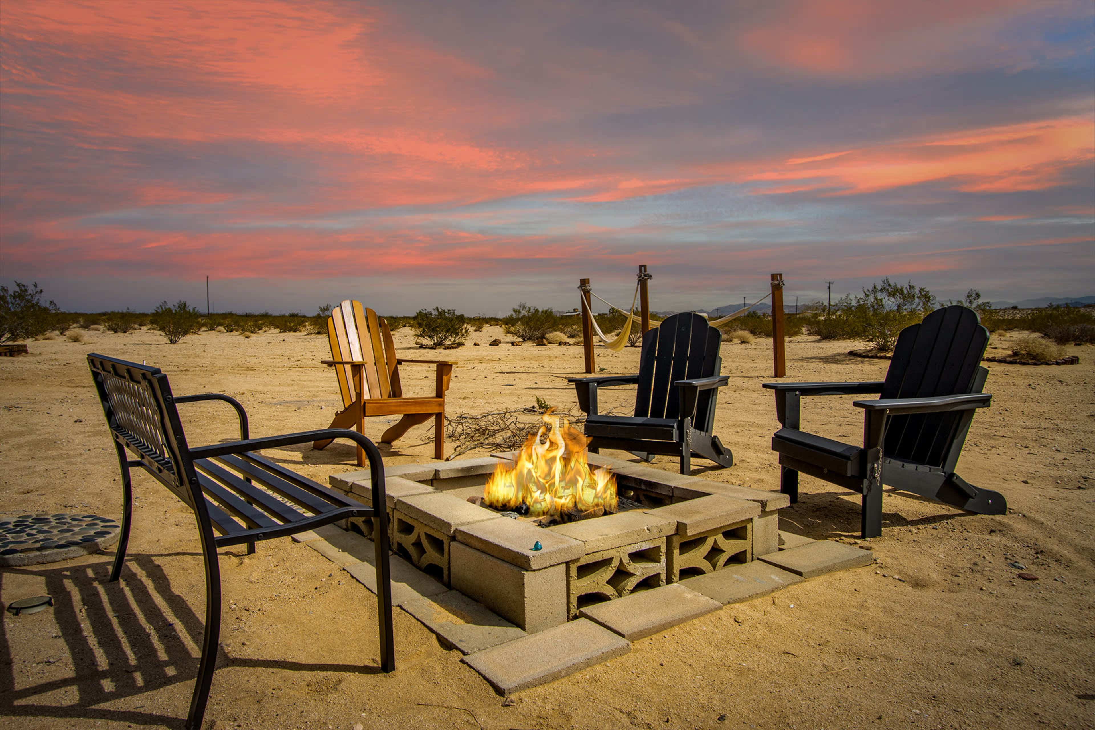 A circular fire pit made of cinder blocks is surrounded by several outdoor chairs in a desert landscape under a colorful sunset.