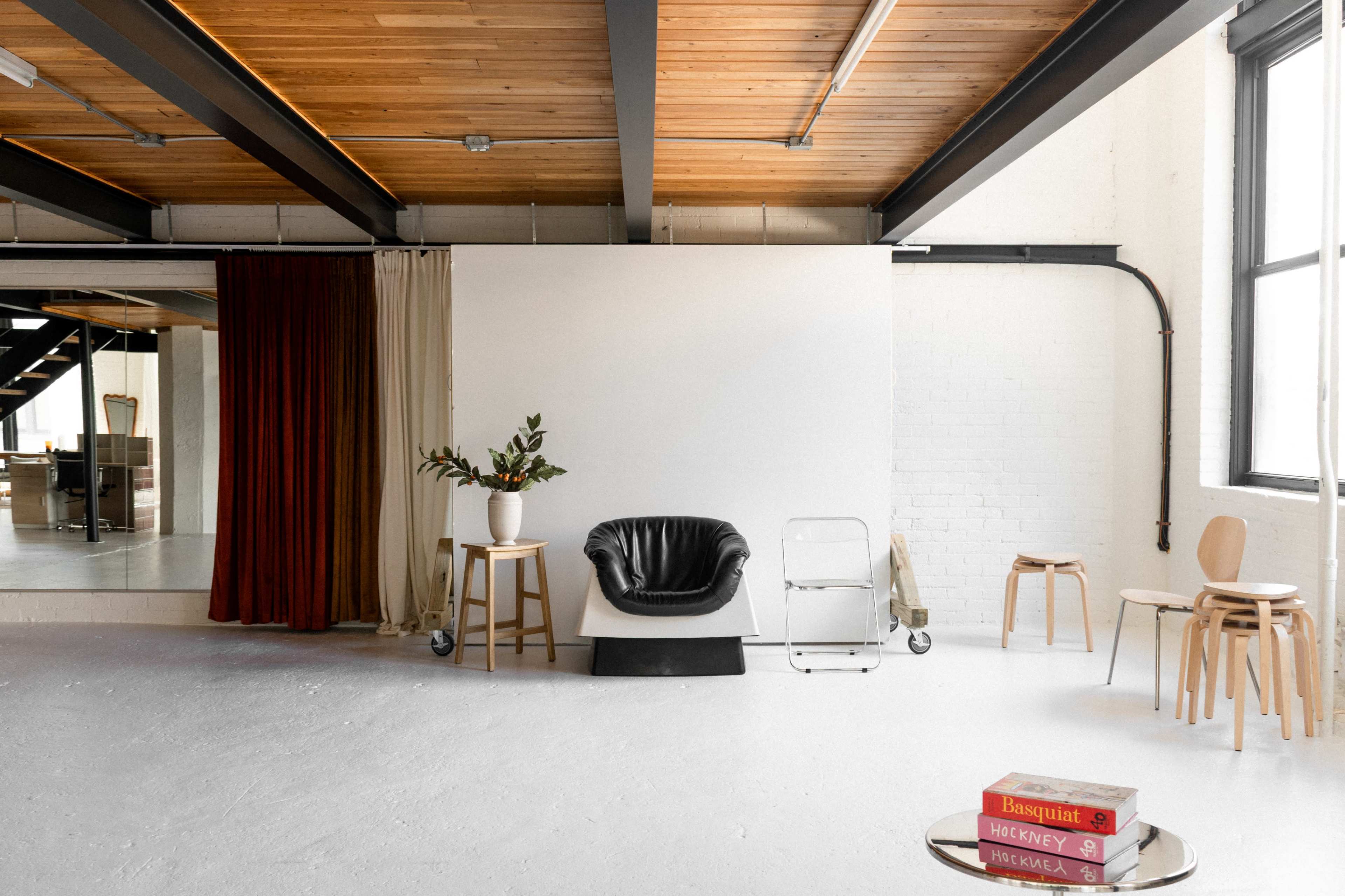 The image shows a minimalist indoor space featuring a black chair, a potted plant on a white table, and wooden stools, with large windows and a wooden ceiling.
