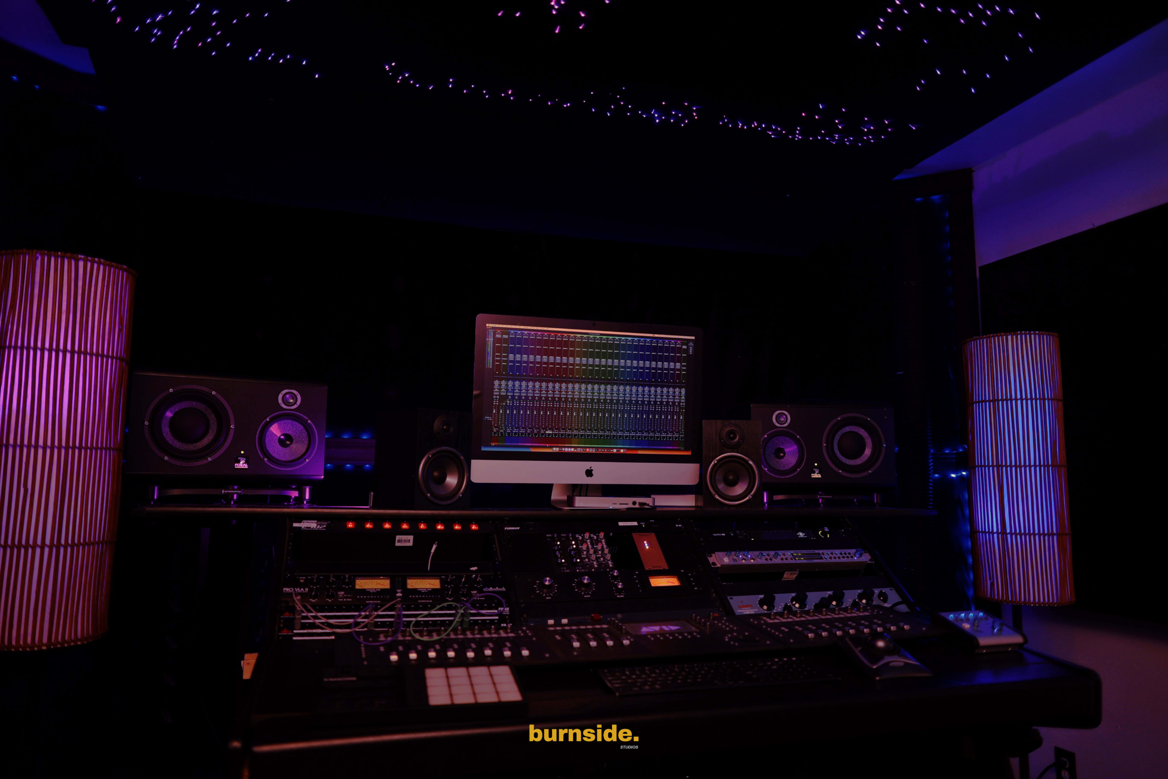 A music studio is shown, featuring a large mixing console, speakers, and an illuminated computer monitor, with decorative lighting in the background.