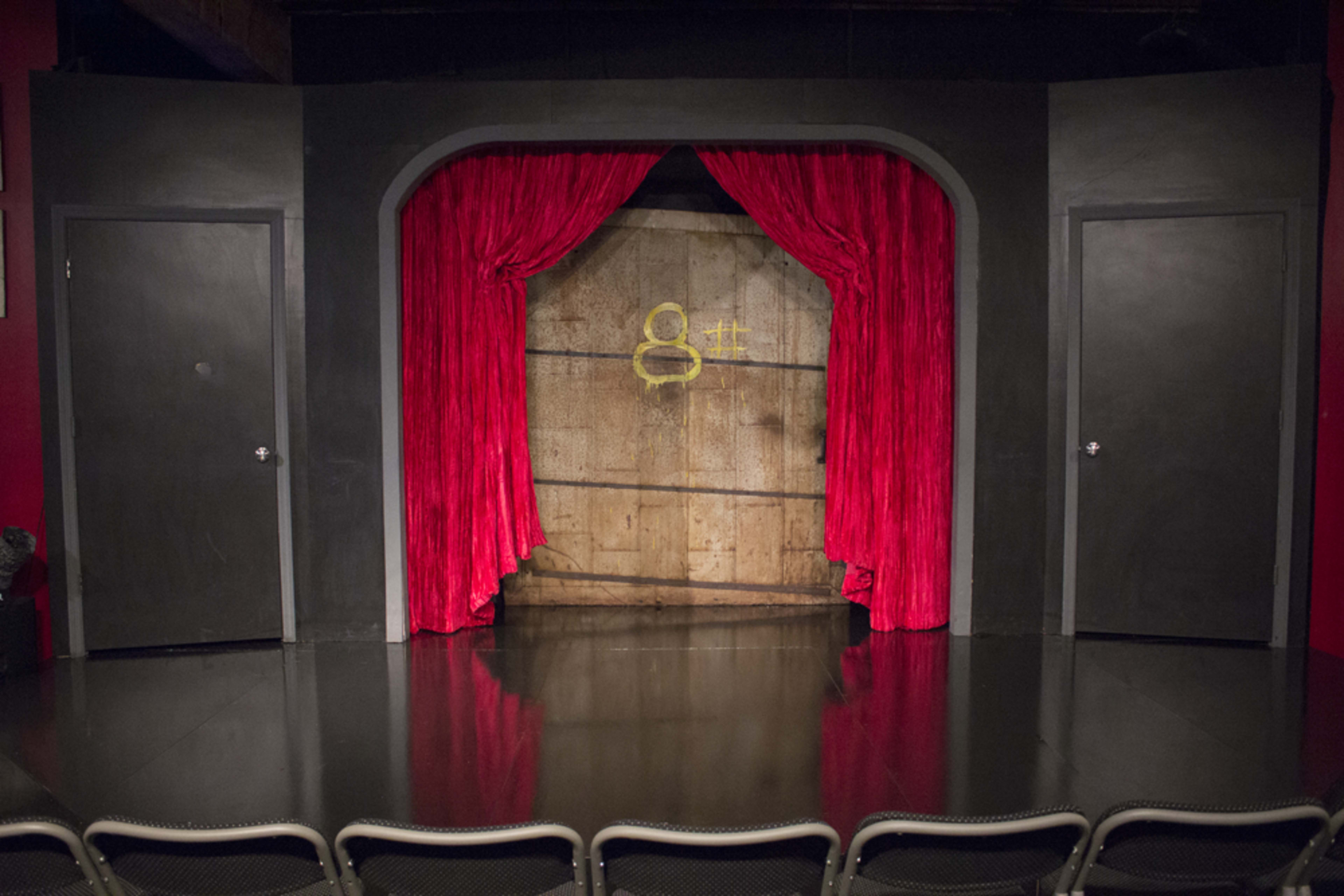 A theater stage with a red curtain and two dark doors on either side, set against a wooden backdrop.
