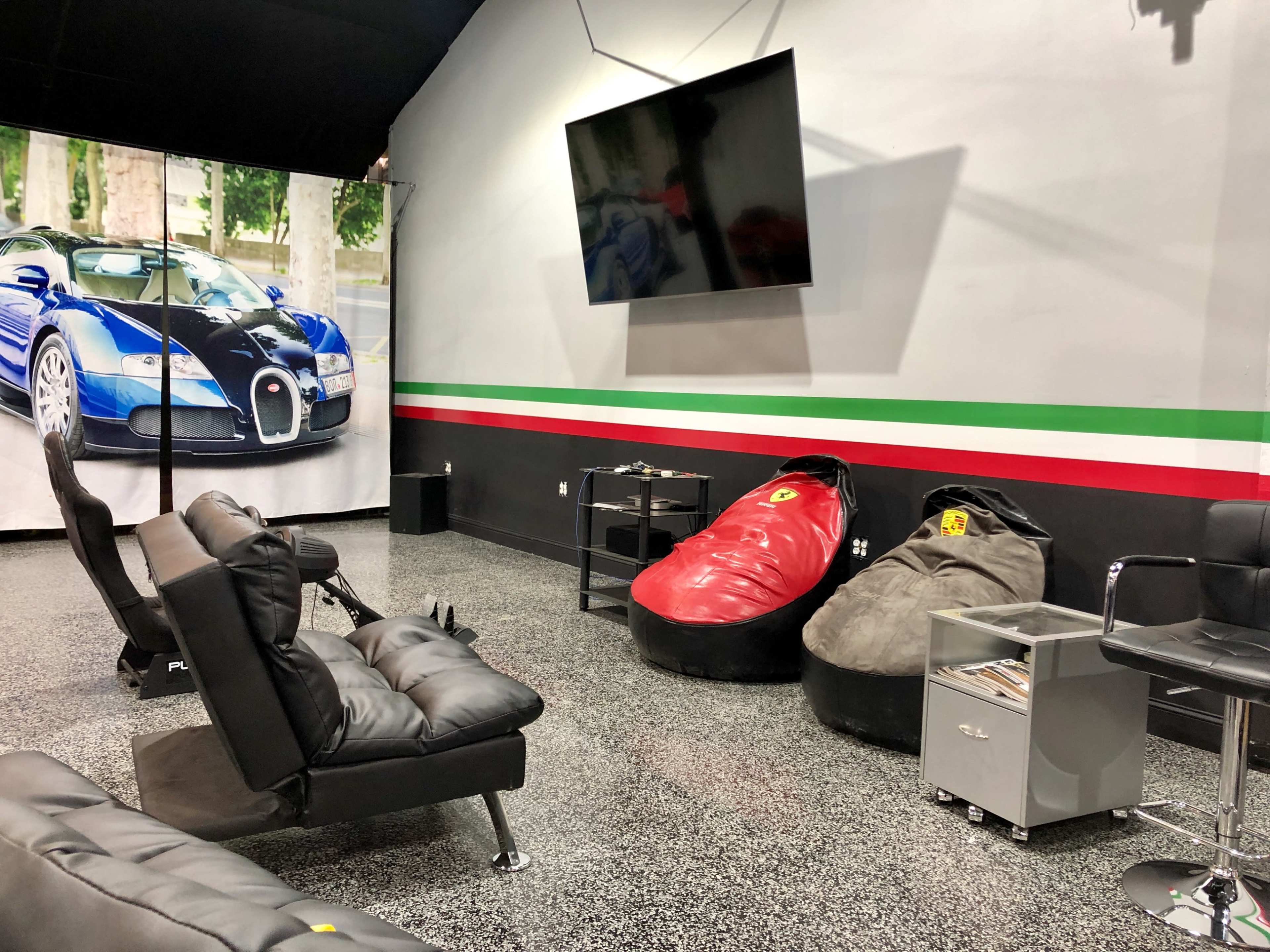 A modern lounge area featuring black leather seating, bean bags resembling sports cars, and a large flat-screen TV mounted on the wall, with a backdrop of a luxury car poster.