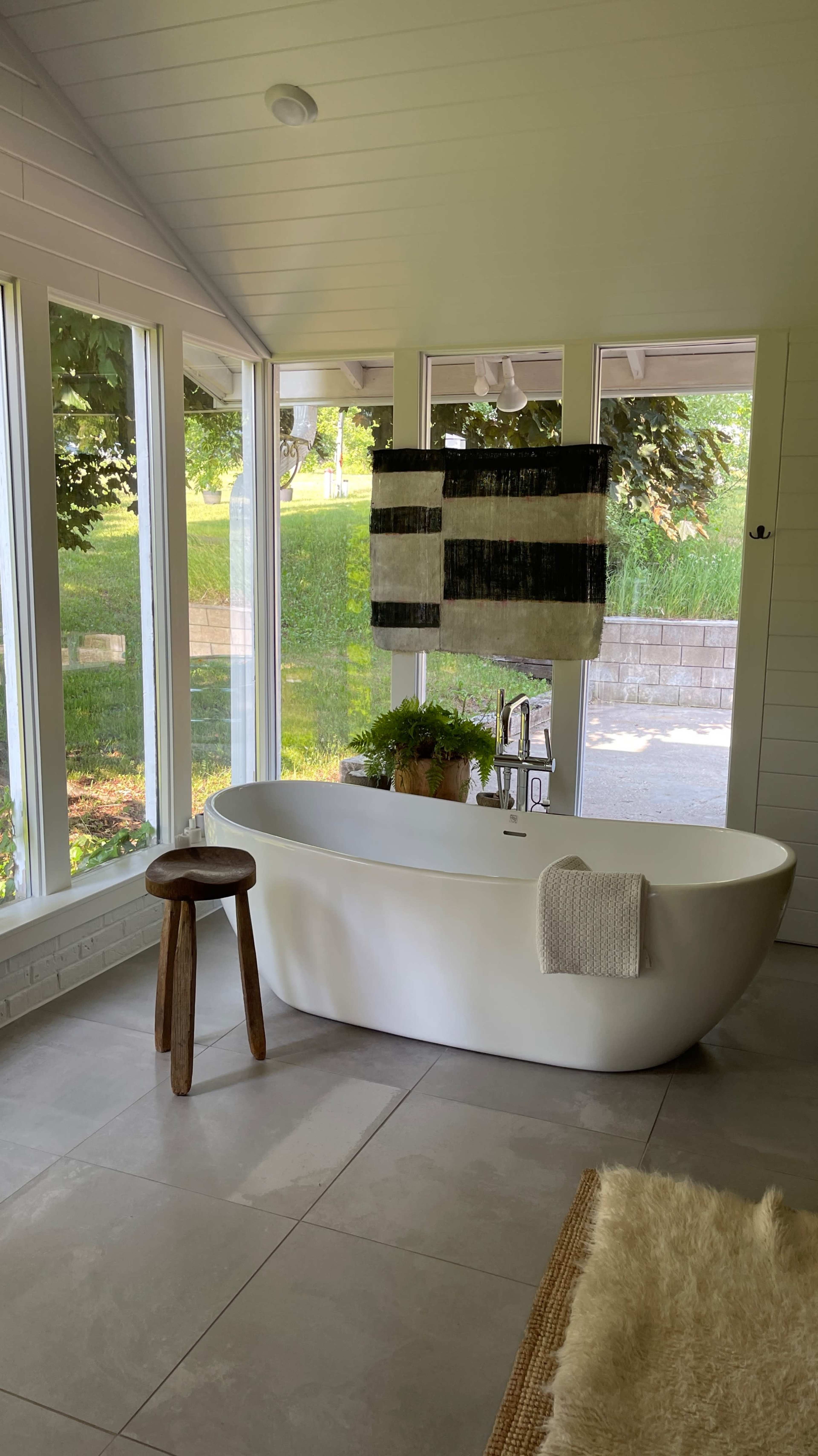 A modern freestanding bathtub is positioned in a brightly lit room with large windows, accompanied by a small stool and a wall hanging.