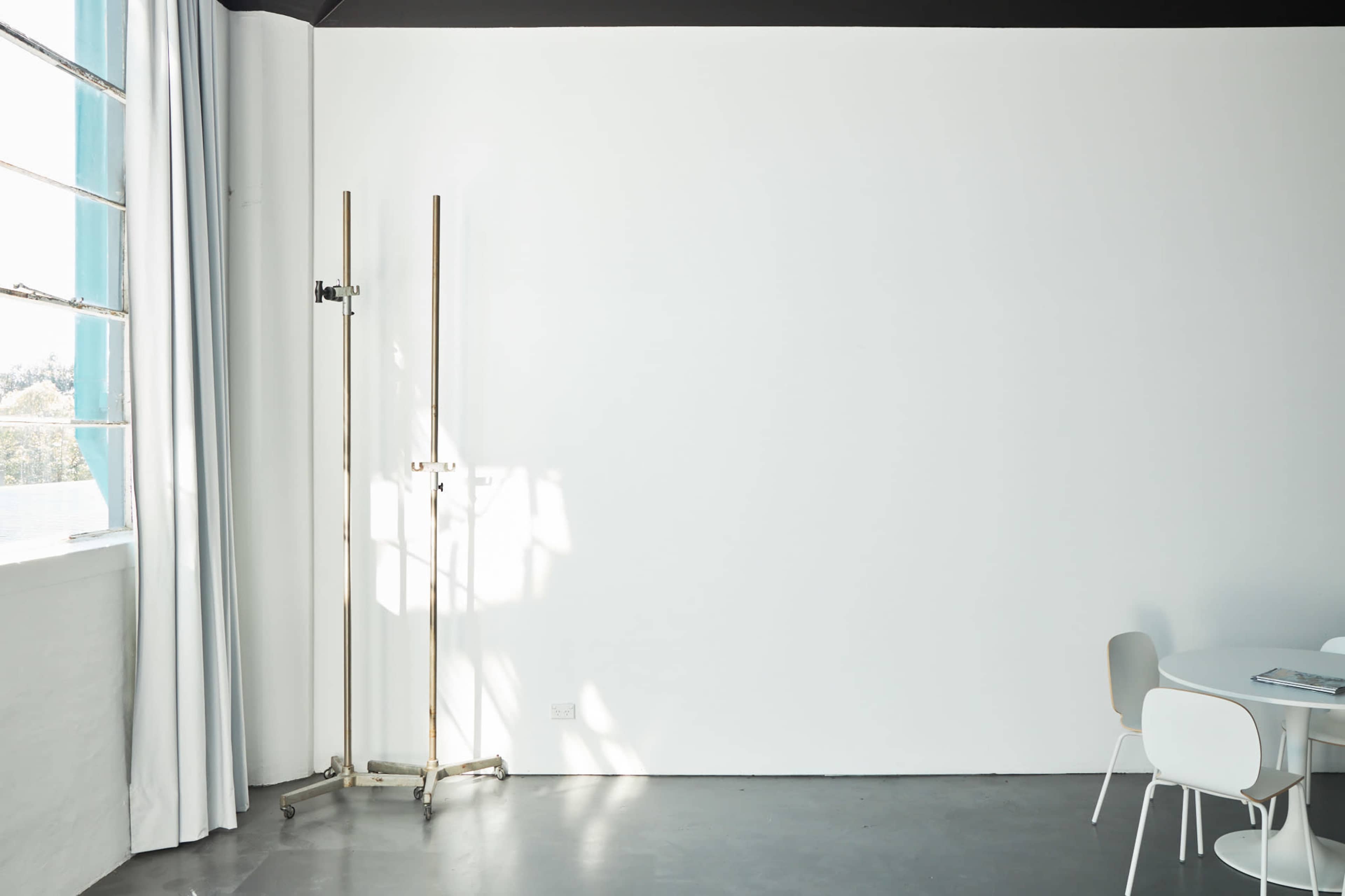 A minimalist room features a blank white wall, a draped curtain, a stand, and a small dining area with simple chairs and a table.
