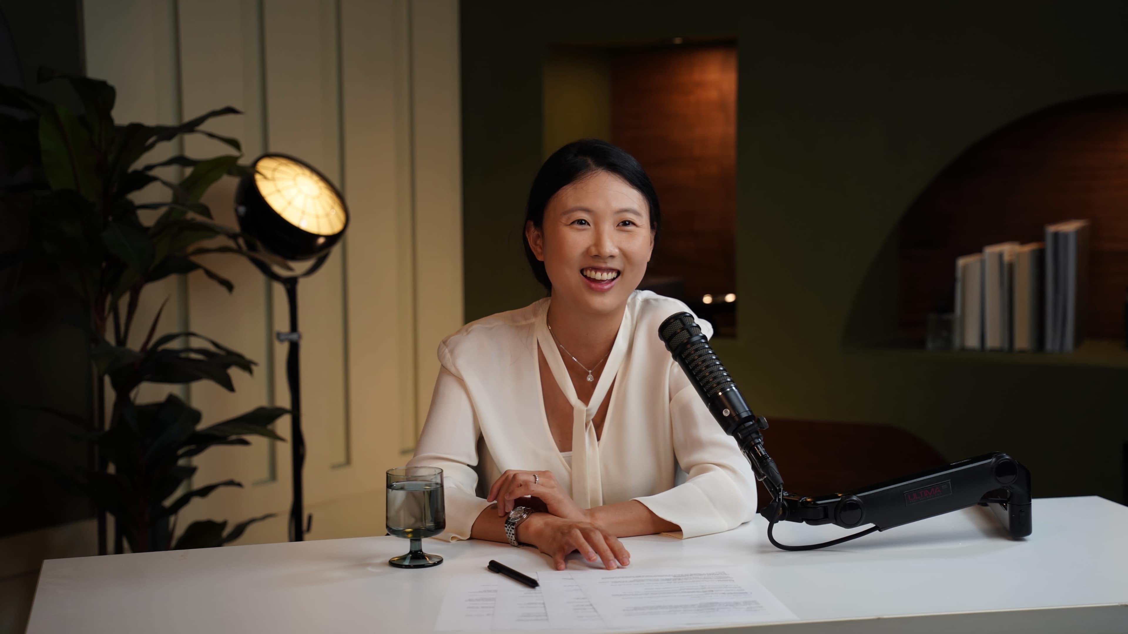 A woman sits at a desk with a microphone and a glass of water, smiling while preparing for a recording session in a well-lit room.