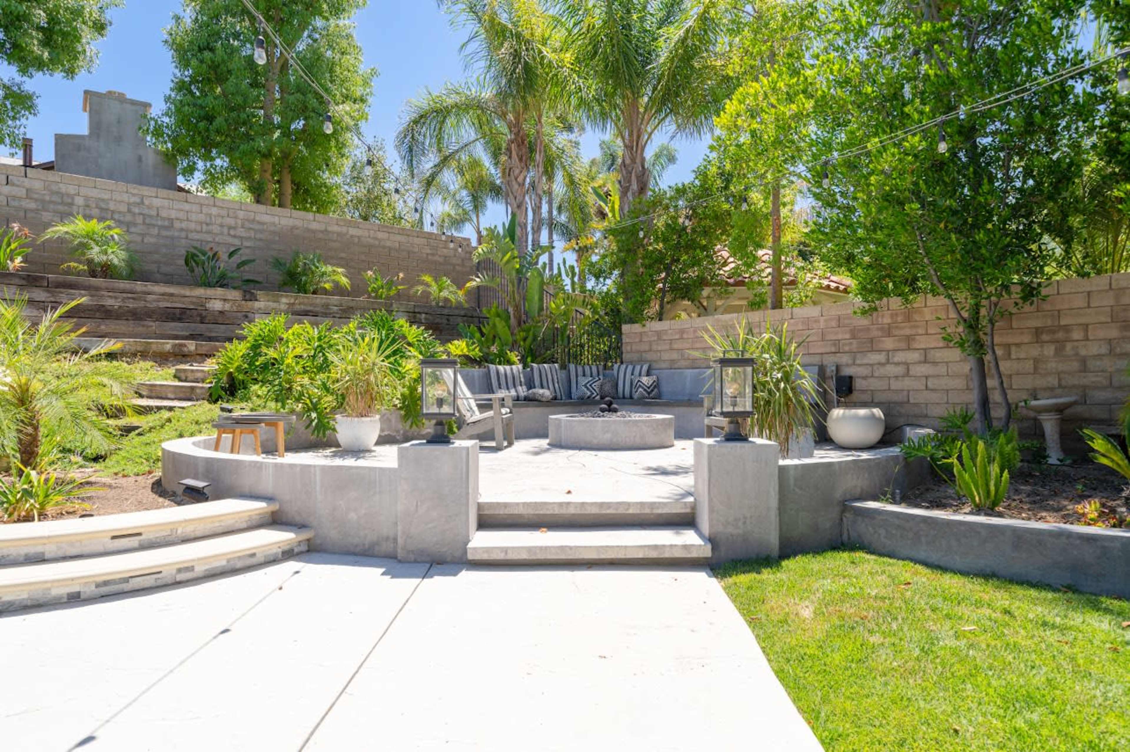A landscaped outdoor seating area with stone steps, a circular fire pit surrounded by chairs, and greenery in the background.