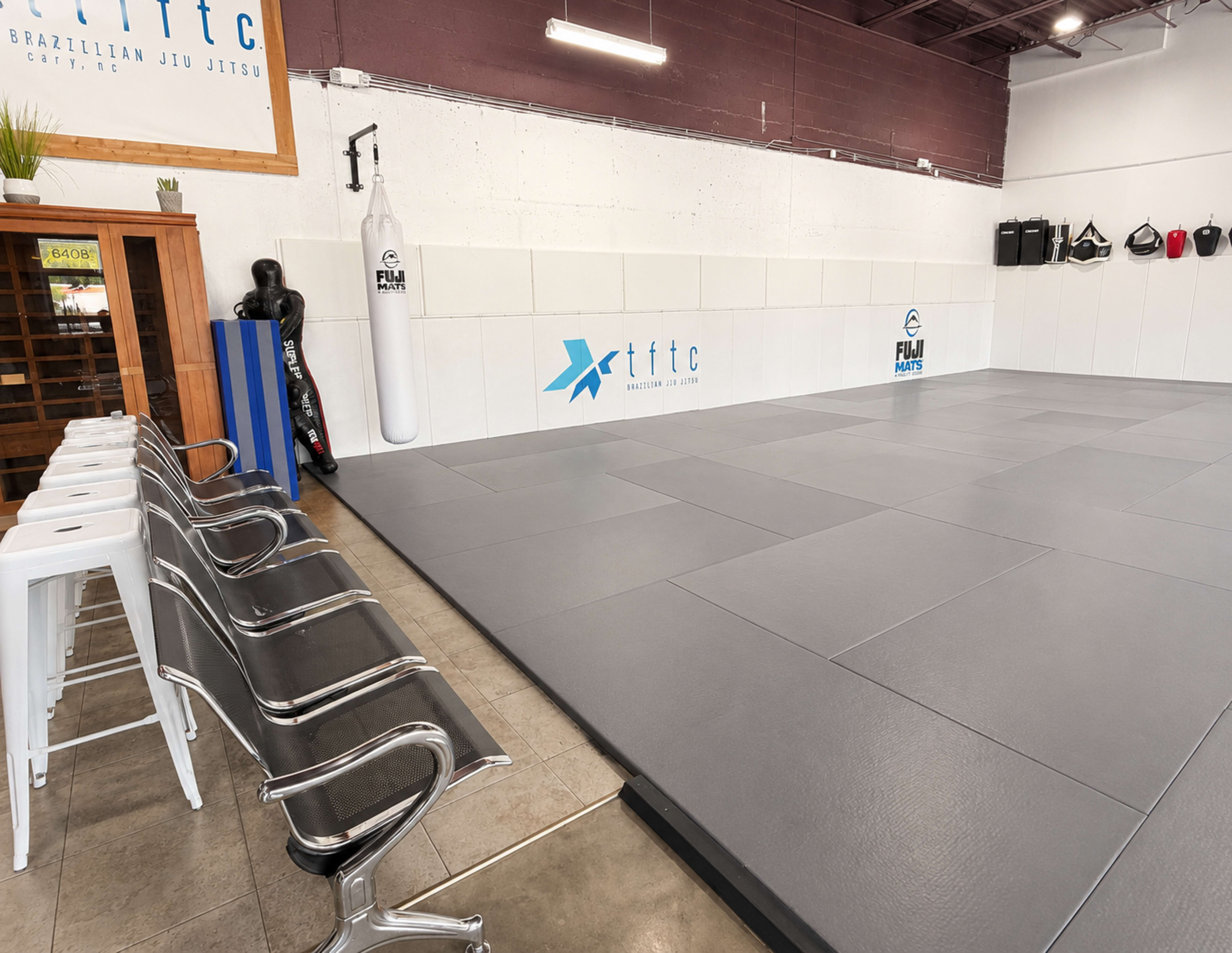 The image shows an empty Brazilian Jiu Jitsu training space with gray mats, a wooden entryway, and equipment hanging on the wall.
