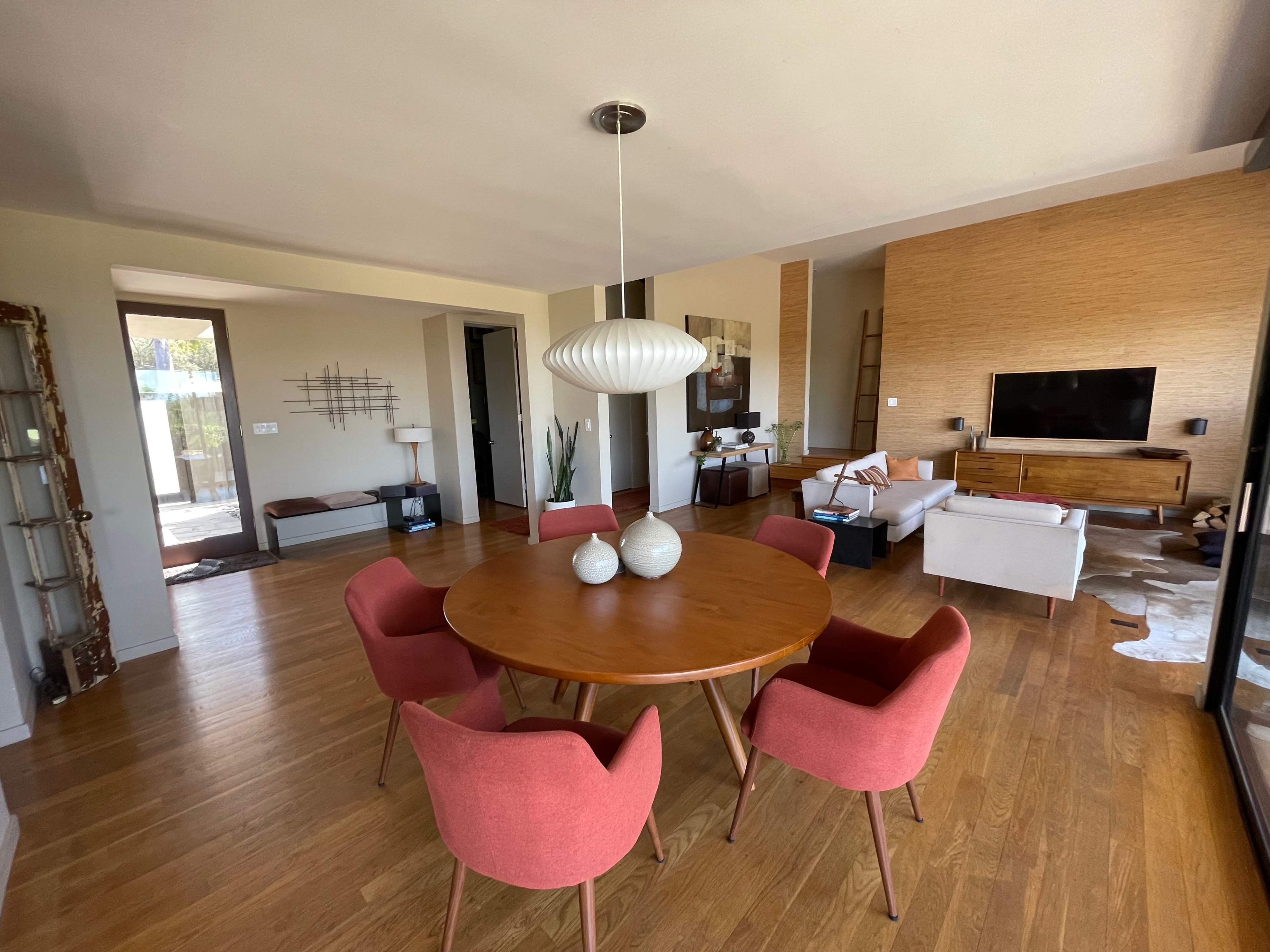 A modern dining area features a round wooden table surrounded by pink chairs, with a living room visible in the background that contains a television and minimalist decor.