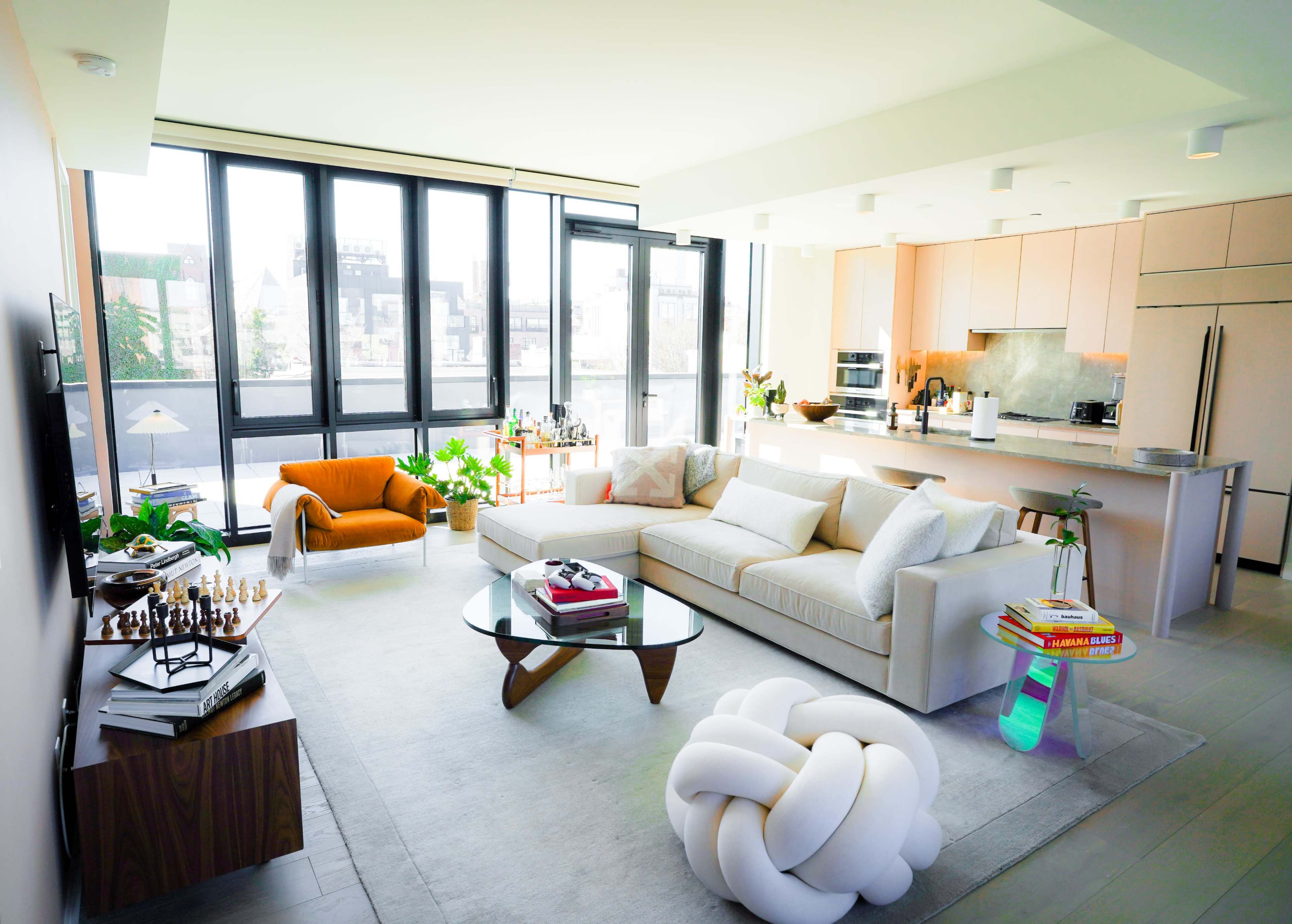 A modern living room with large windows, a sectional couch, a coffee table, and an open kitchen area featuring light-colored cabinetry.
