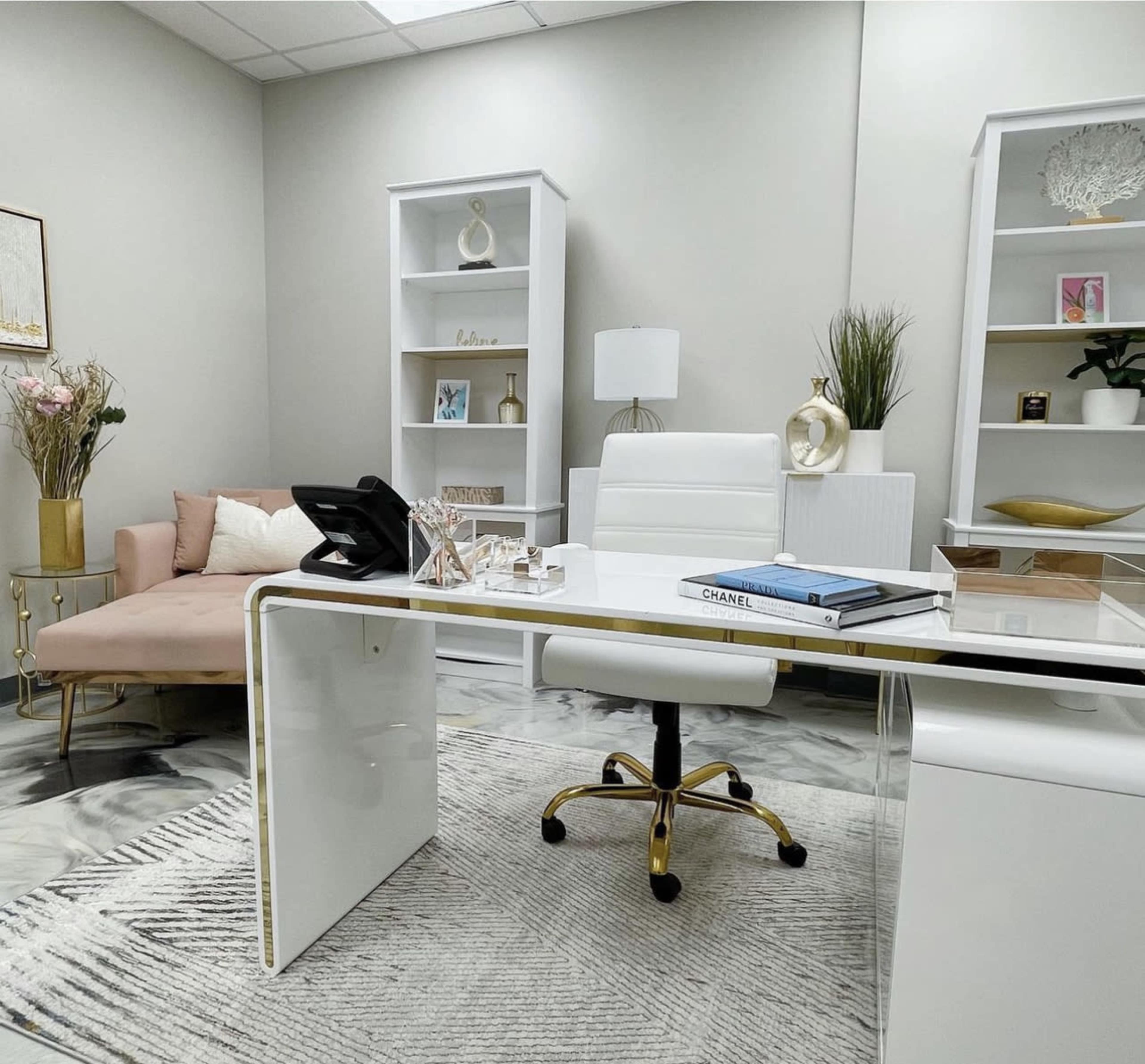 A modern office space featuring a white desk with a gold accent, a white chair, a phone, and shelves containing decorative items, alongside a pink couch and plants.