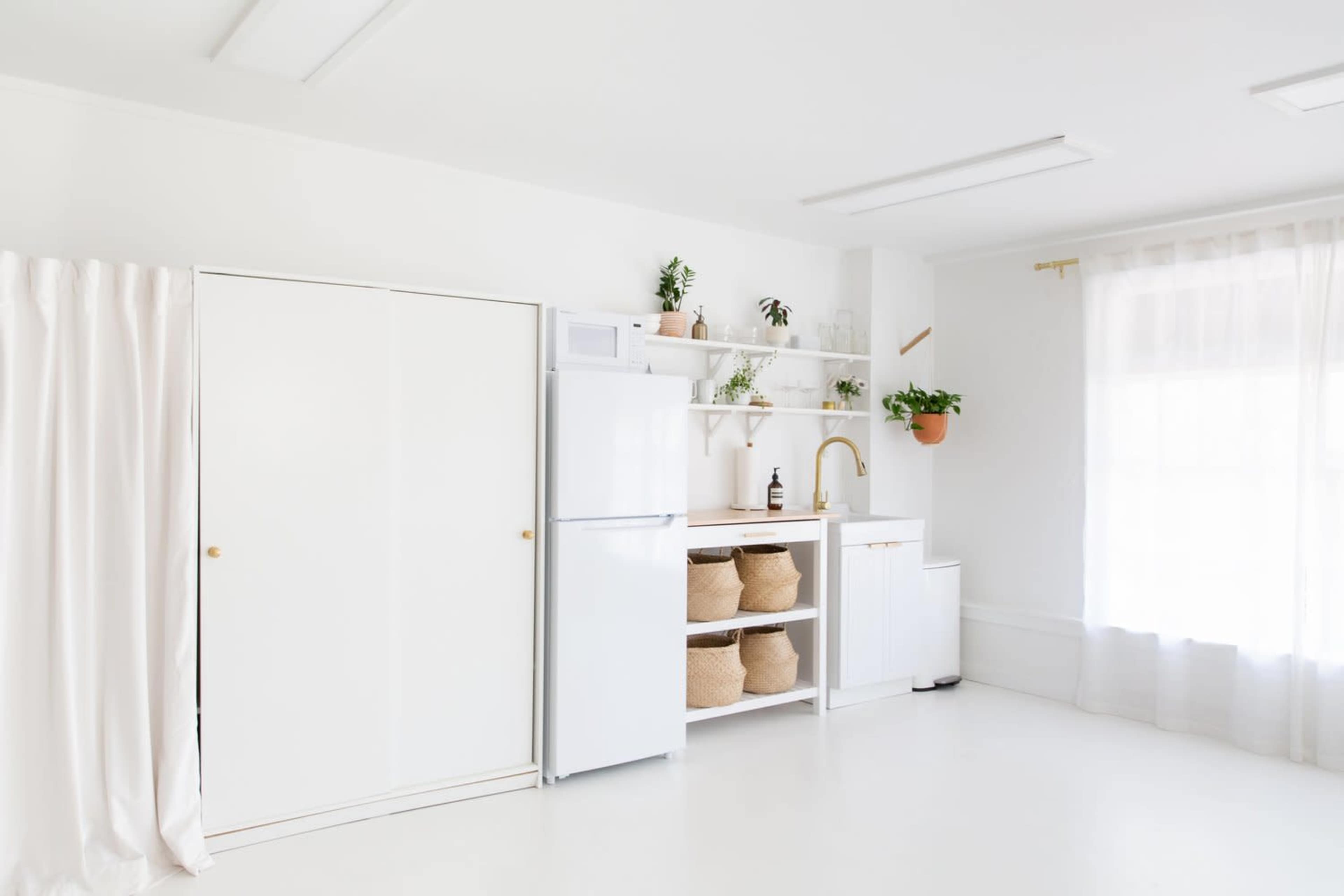 A modern kitchen features white cabinetry, a refrigerator, open shelving with plants, and natural light streaming through large windows.