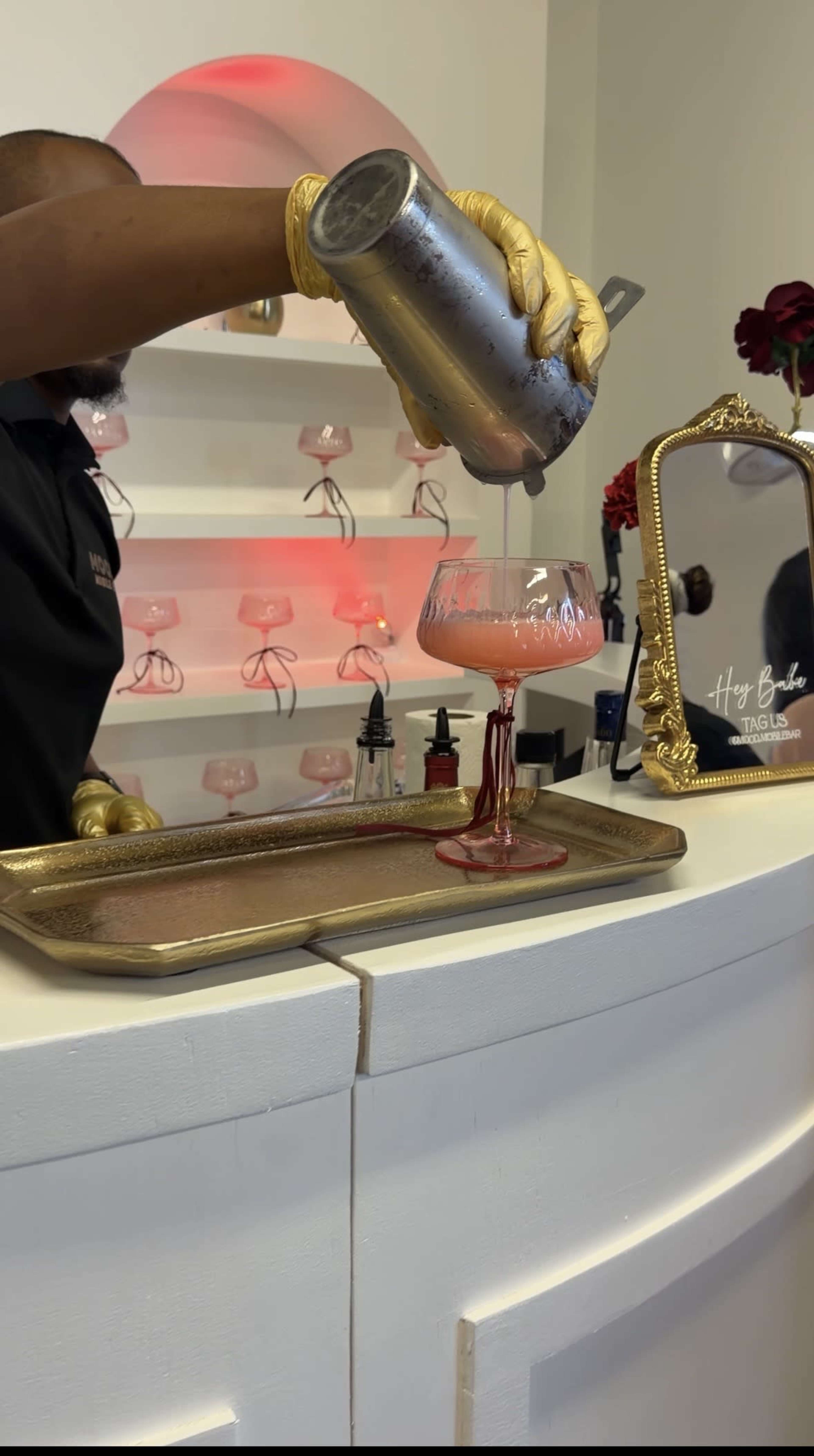A bartender wearing gloves pours a pink cocktail from a shaker into a glass on a gold tray in a stylish bar setting.