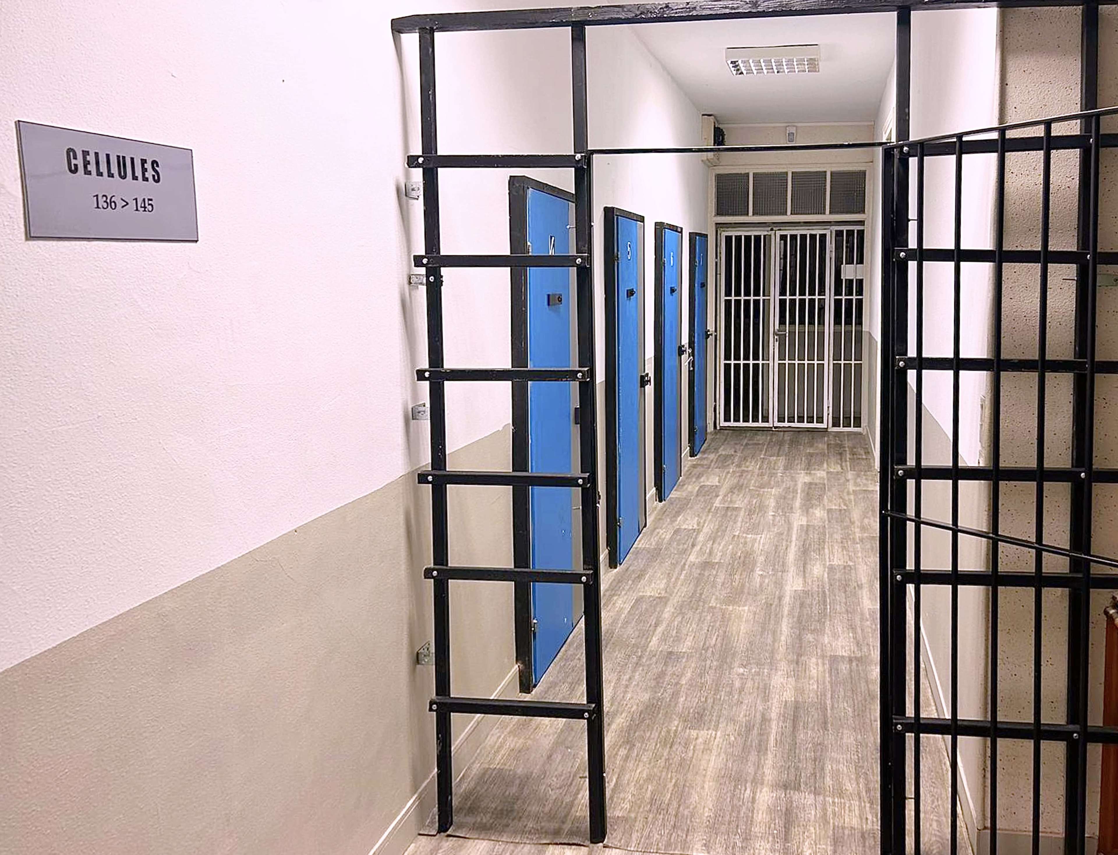 A narrow hallway in a prison with blue cell doors and a barred gate at the entrance.
