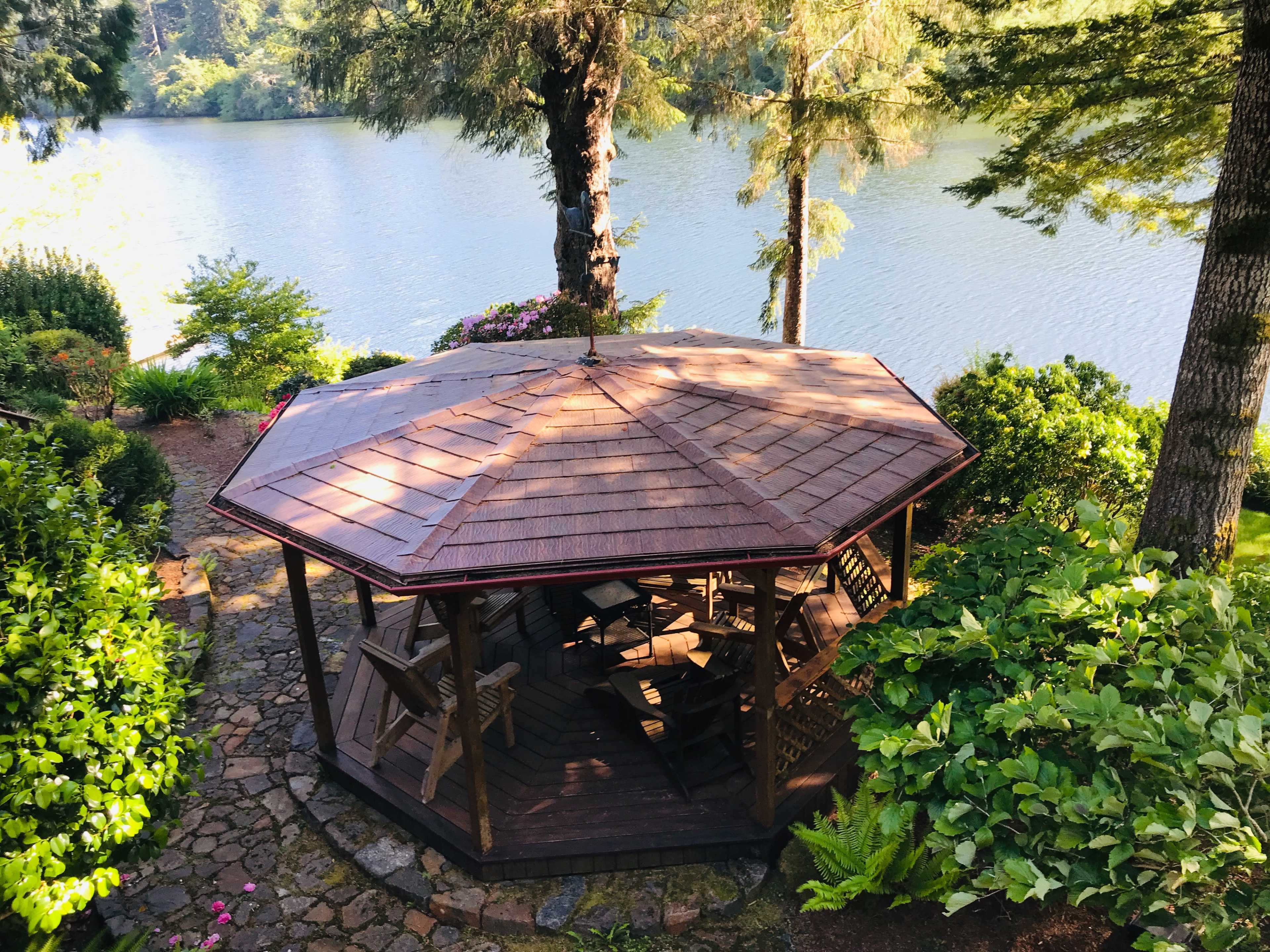 A wooden octagonal gazebo is situated beside a calm lake, surrounded by greenery and blooming flowers.