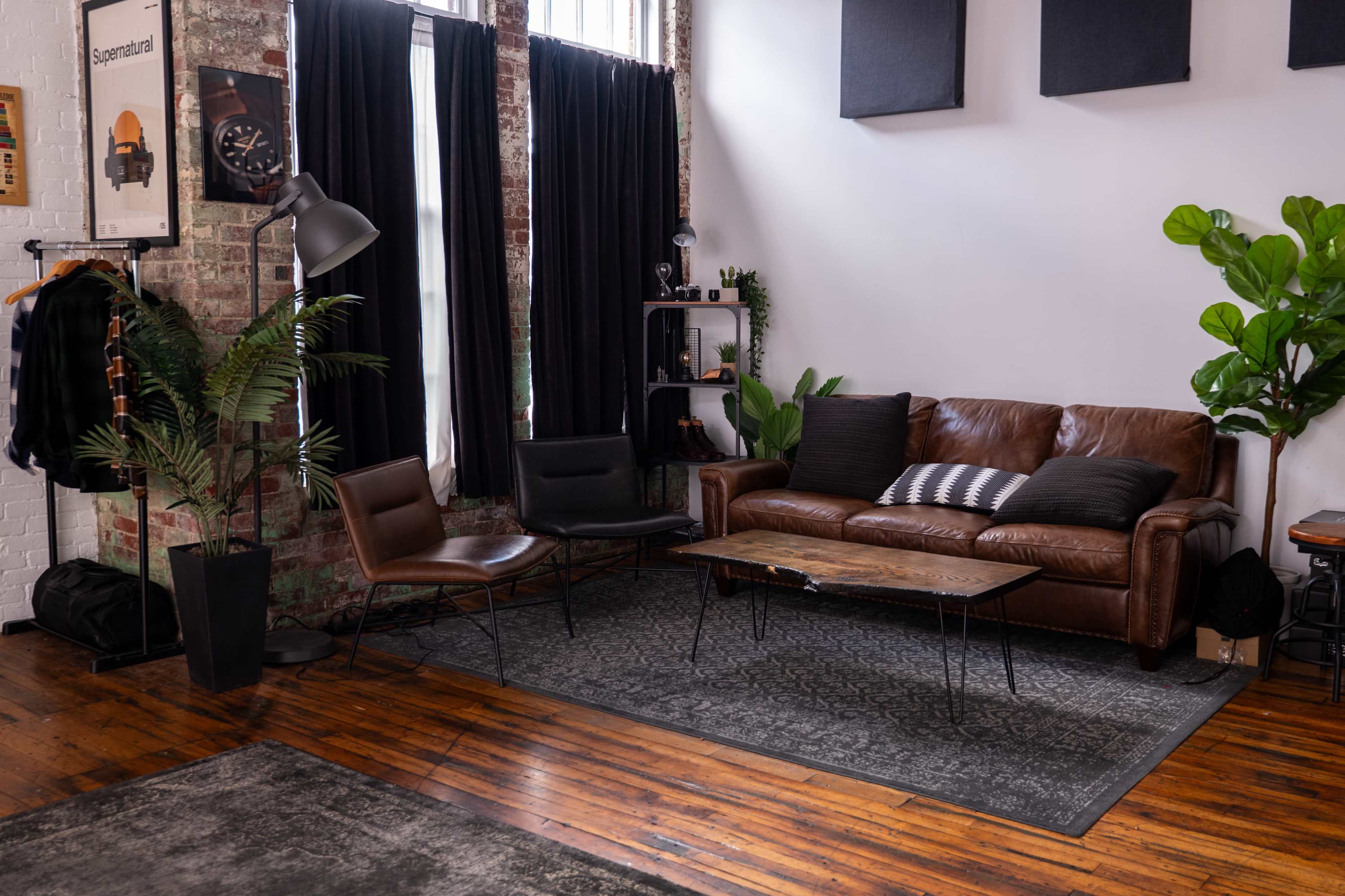 The image shows a cozy living space featuring a brown leather sofa, two dark chairs, a wooden coffee table, and plants, set against a backdrop of exposed brick walls and large windows.