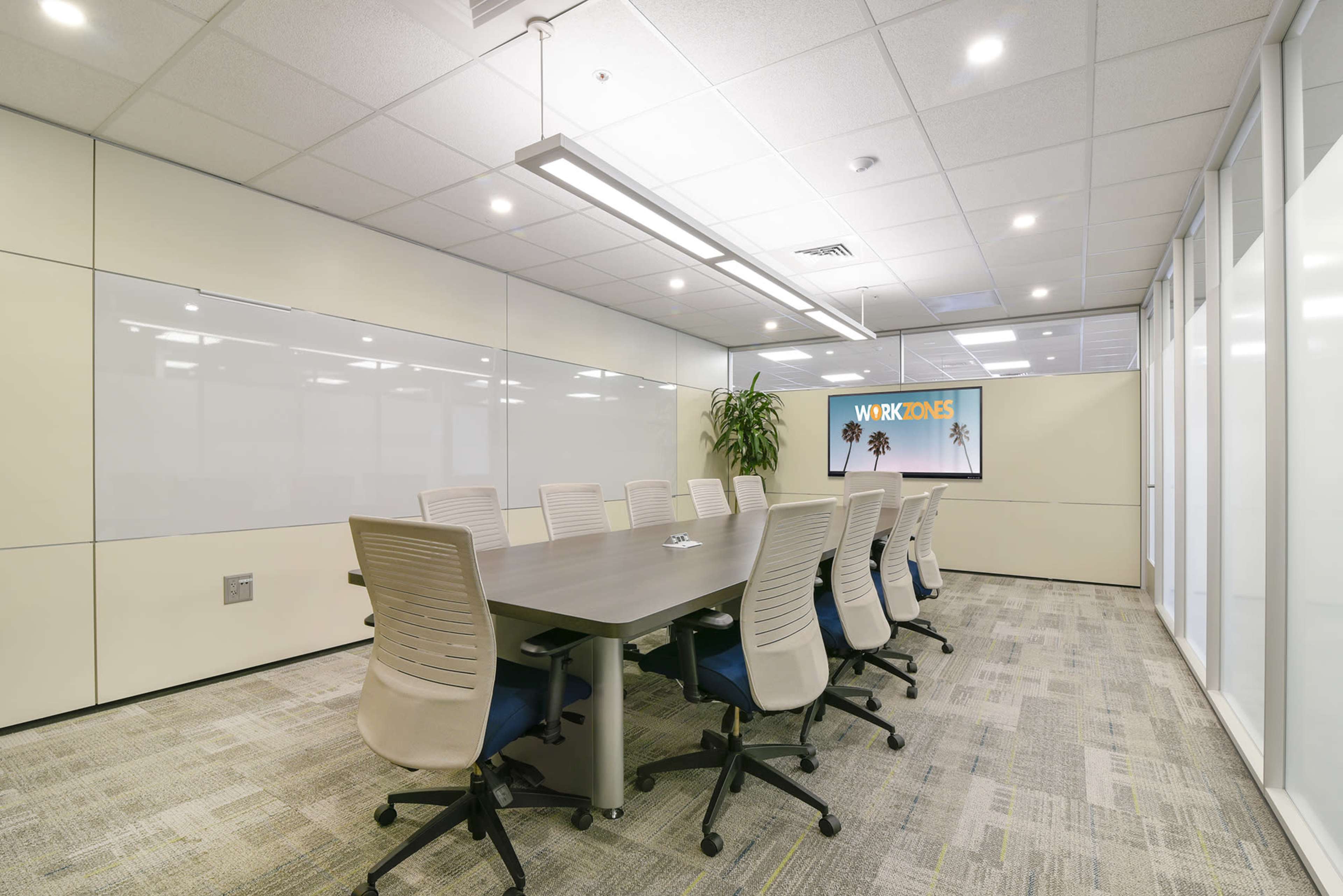 A modern conference room features a long table surrounded by ergonomic chairs, with a large screen displaying "WORKZONES" and glass walls allowing natural light.