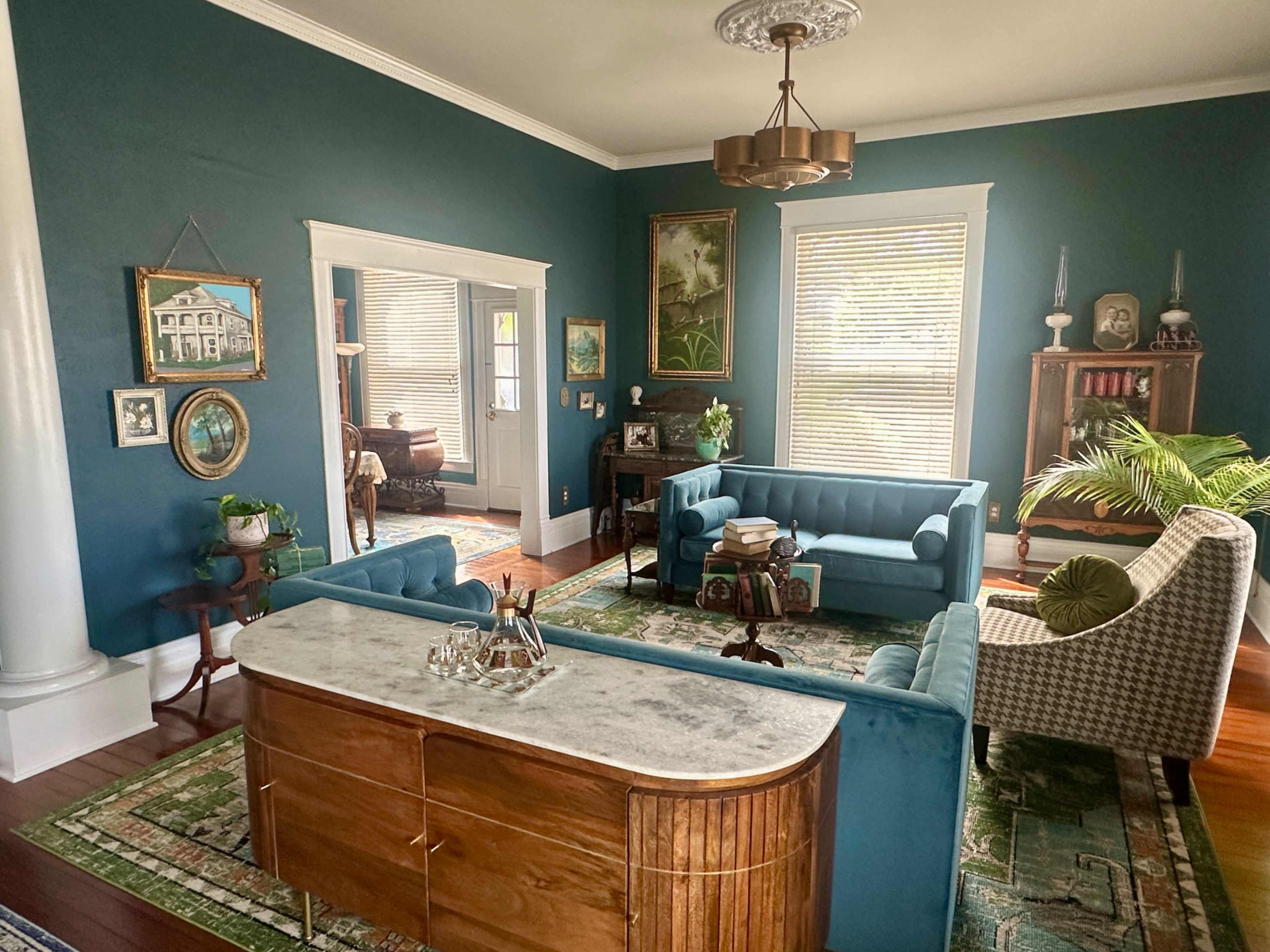 The image shows a brightly lit living room with teal walls, featuring a marble-top sideboard, two teal sofas, and various decorative items.