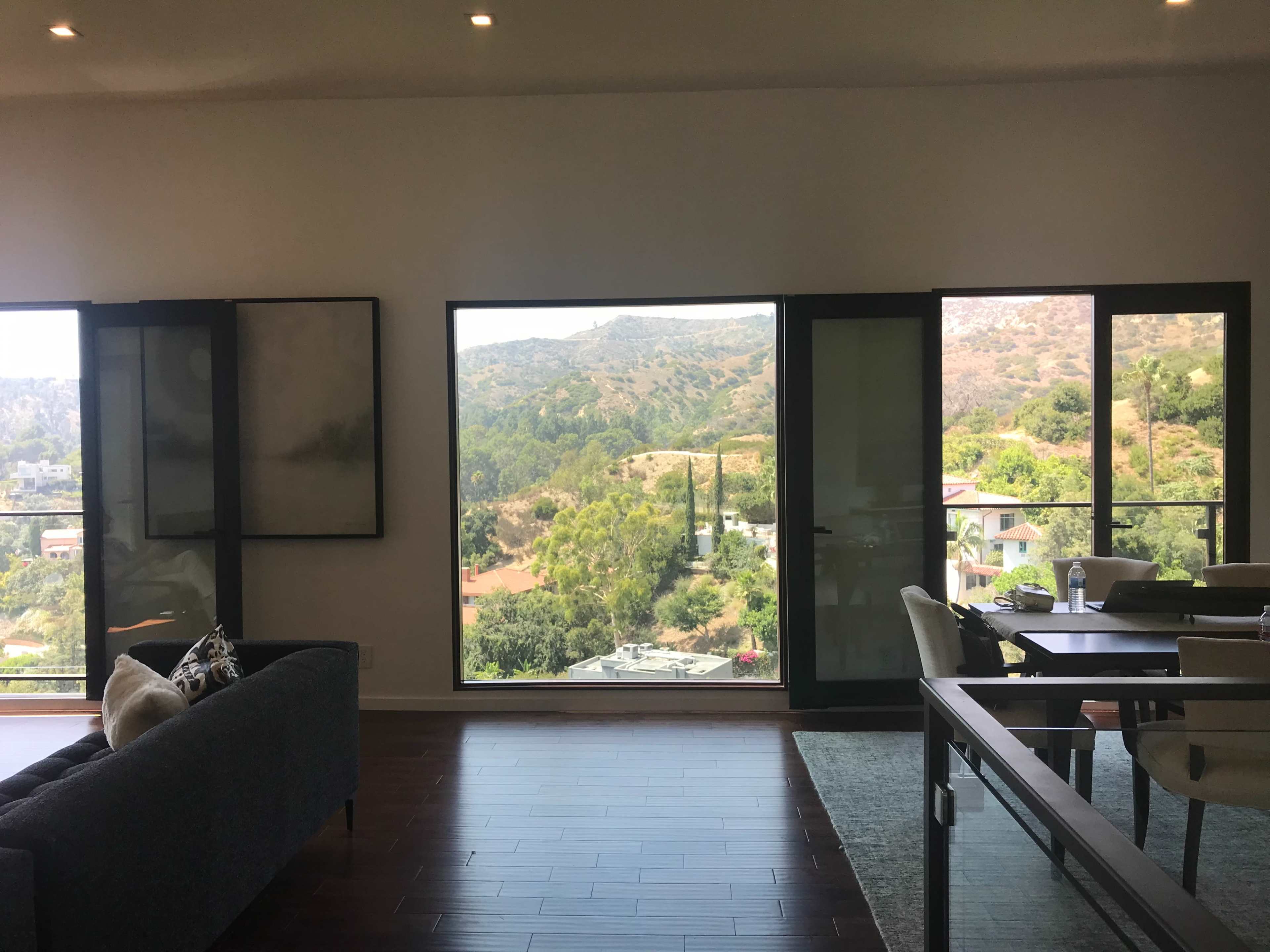 A modern living room with large windows overlooking a green hillside and adjacent buildings.