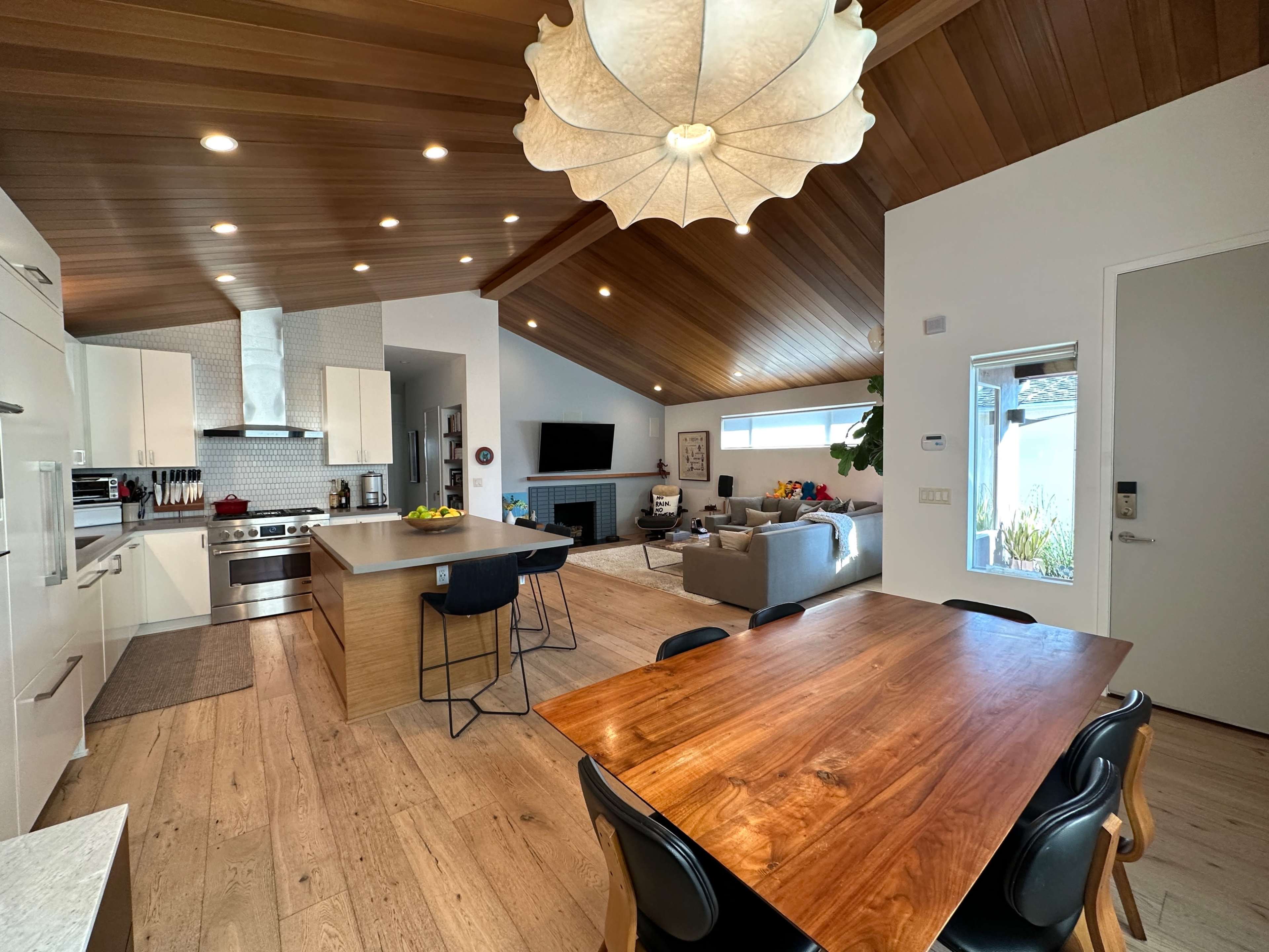 A modern kitchen and dining area feature wooden ceilings, a large central island, and a table surrounded by black chairs.