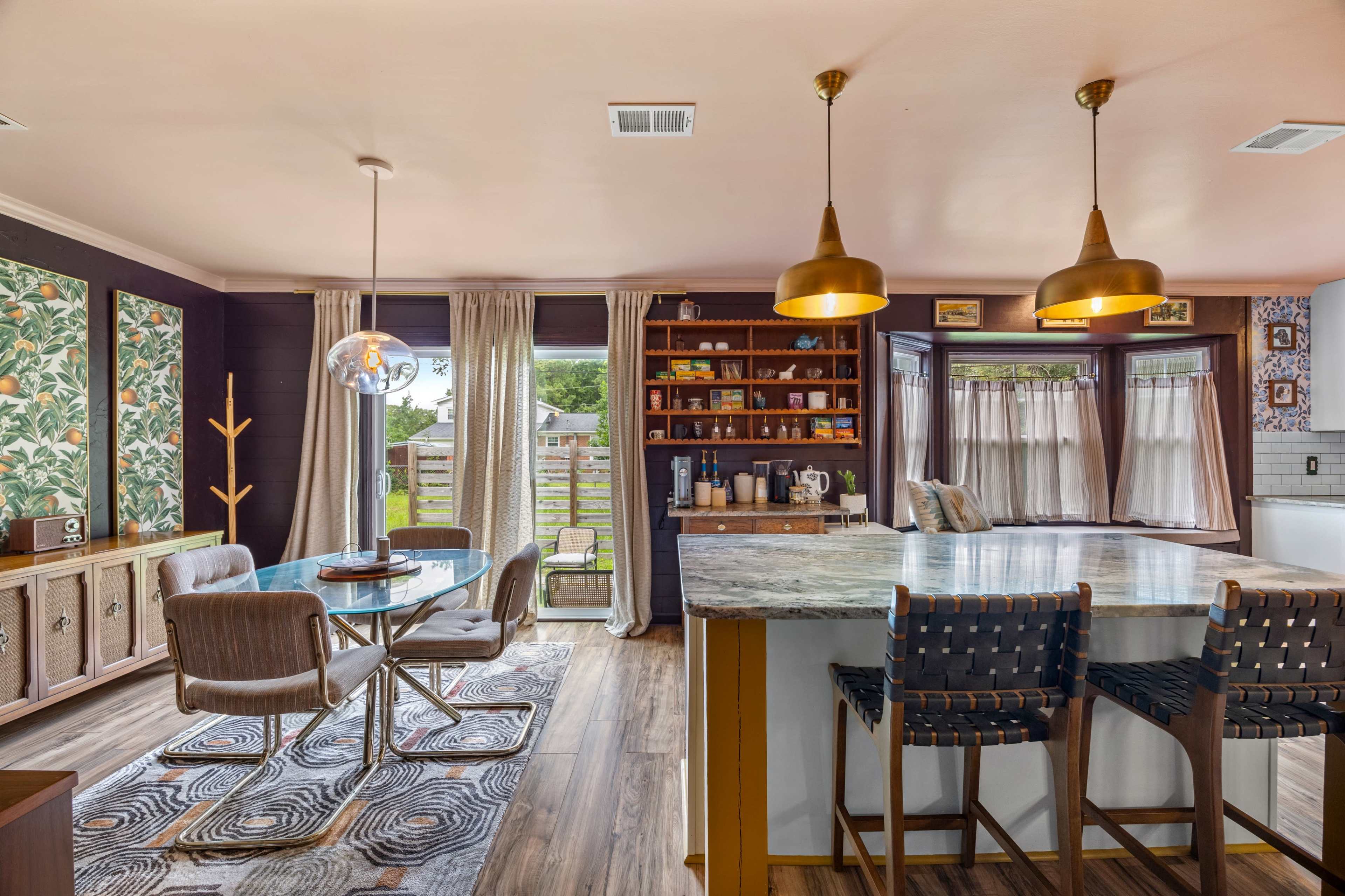 The image shows a modern kitchen and dining area with a circular dining table, a kitchen island with bar stools, and large windows that provide natural light.