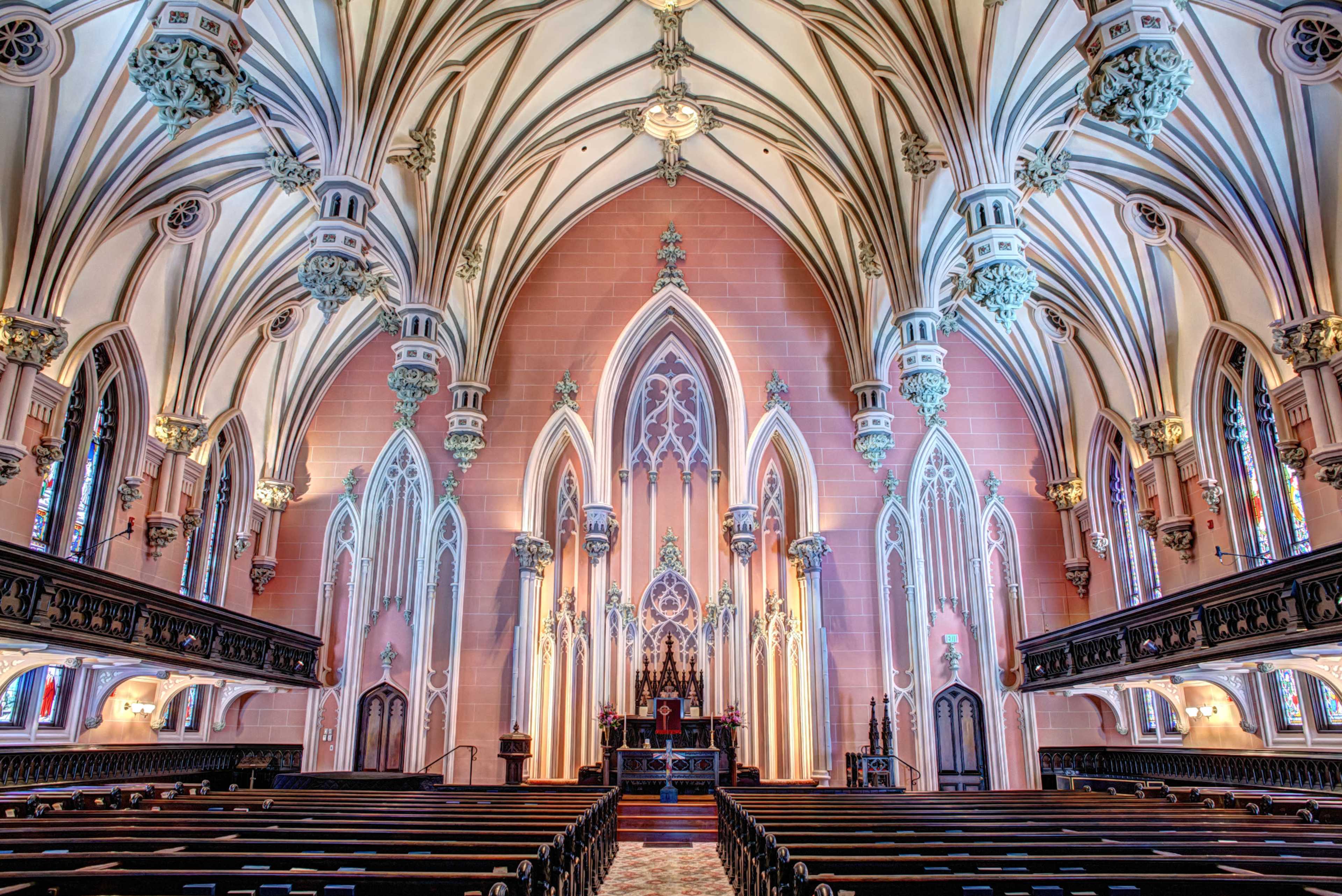 The image shows the interior of a grand church with high vaulted ceilings, intricate architectural details, stained glass windows, and rows of wooden pews facing an ornate altar.