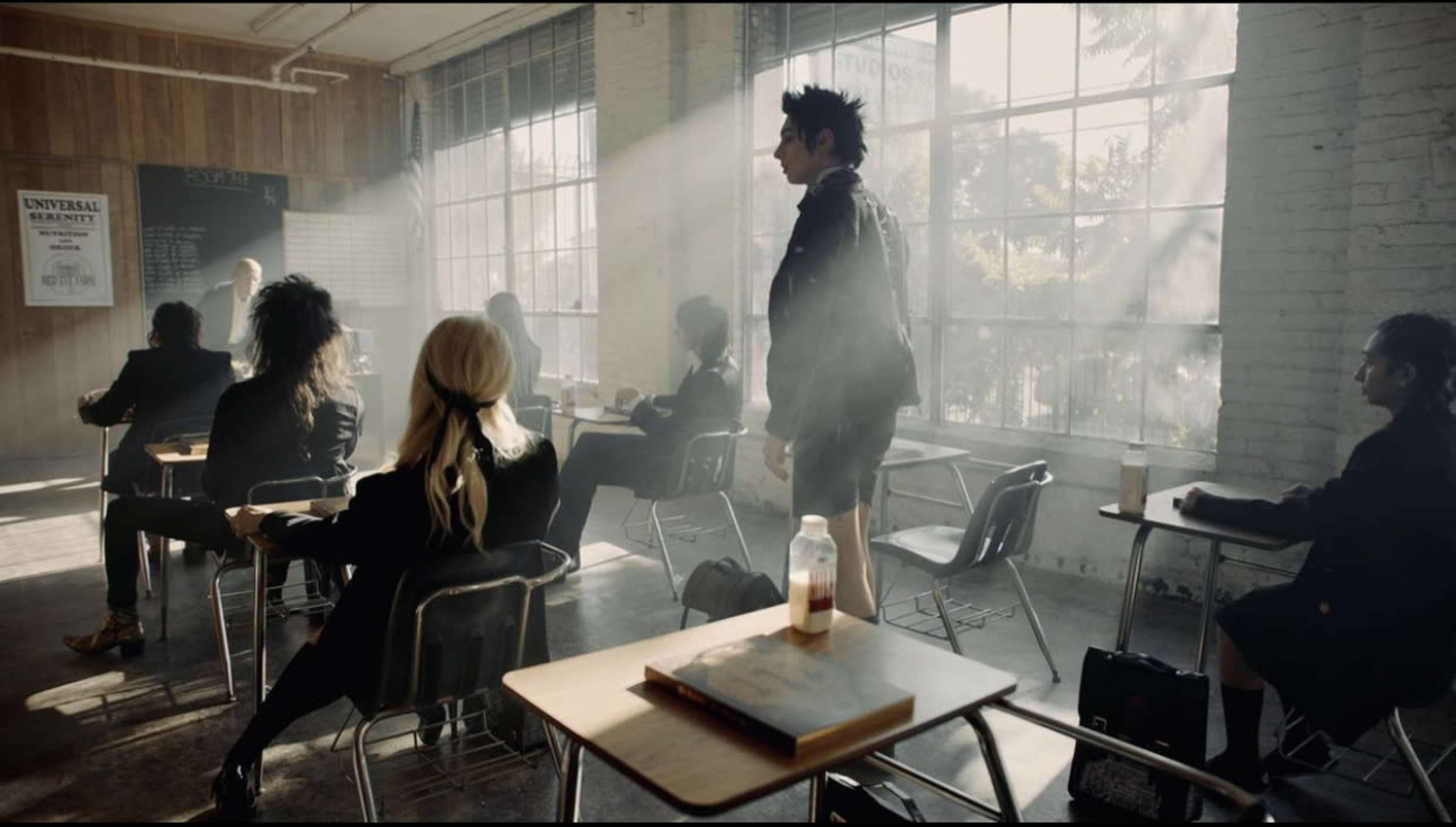 A classroom scene features several students sitting at desks while one student stands, with sunlight streaming through large windows.