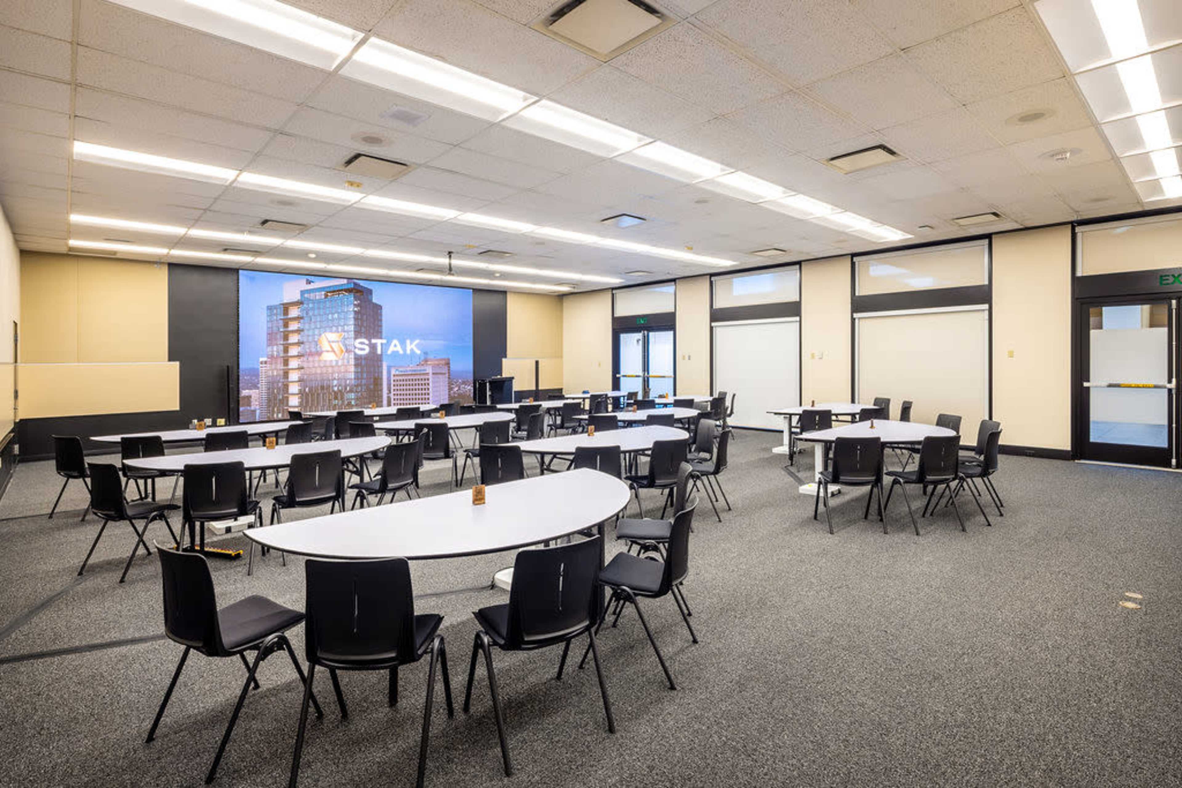 The image shows a spacious, empty conference room with multiple white tables arranged in a U-shape and black chairs around them, featuring a large screen displaying the word "STAK" on one wall.