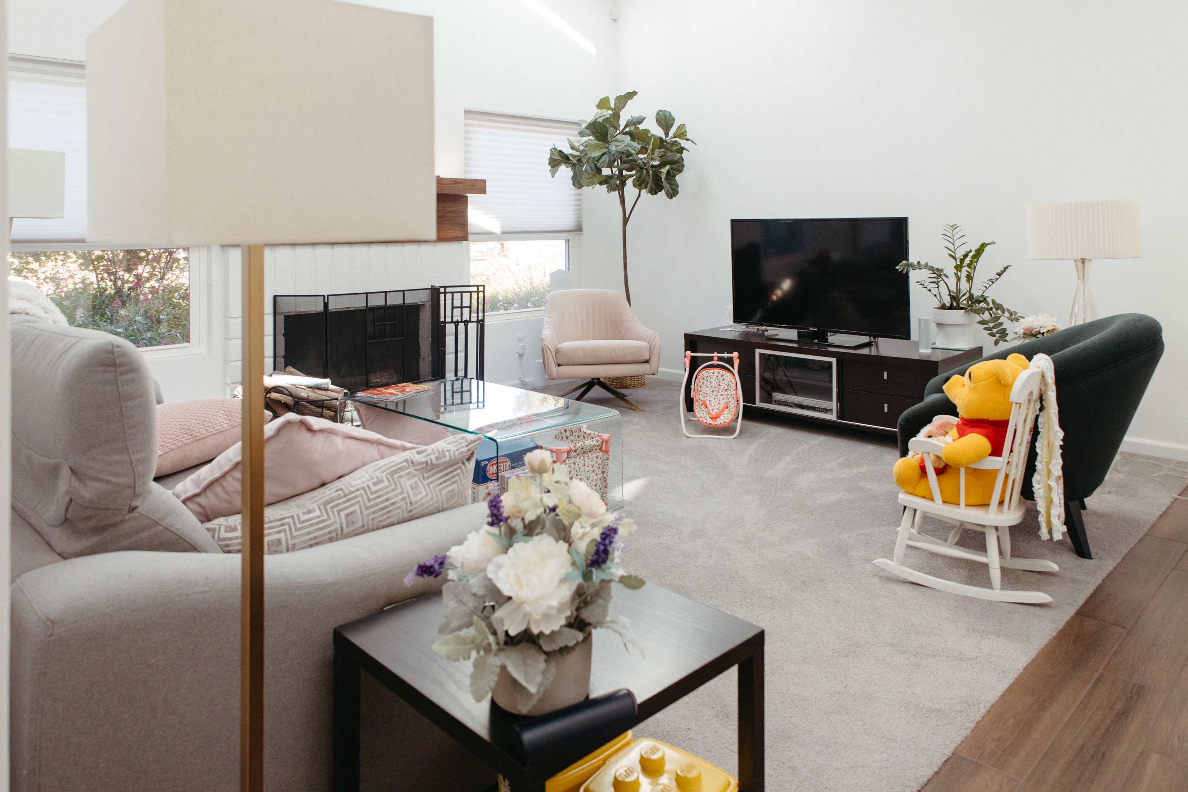The image shows a modern living room with a television, two sofas, a rocking chair, a coffee table, and a potted plant, all arranged on a carpeted floor.
