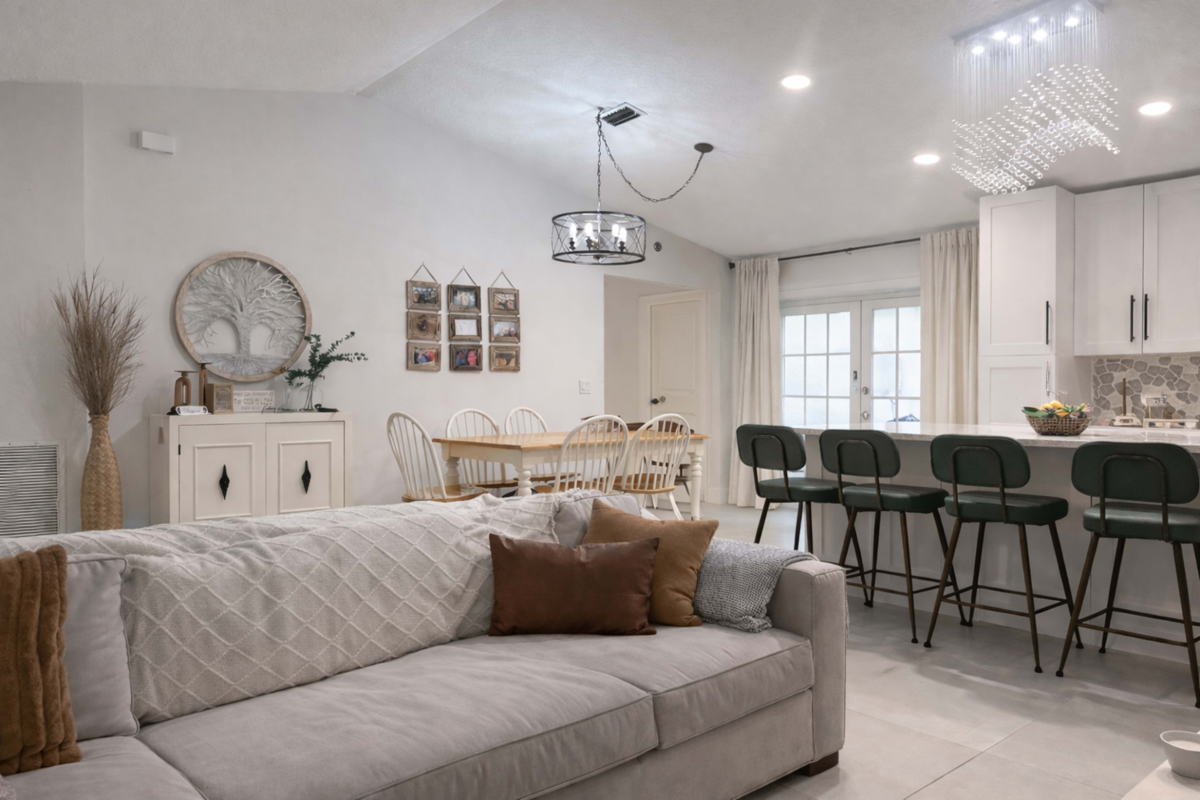 The image shows a modern living room featuring a gray sofa with decorative pillows, a dining area with a wooden table and white chairs, and a kitchen bar with high stools.