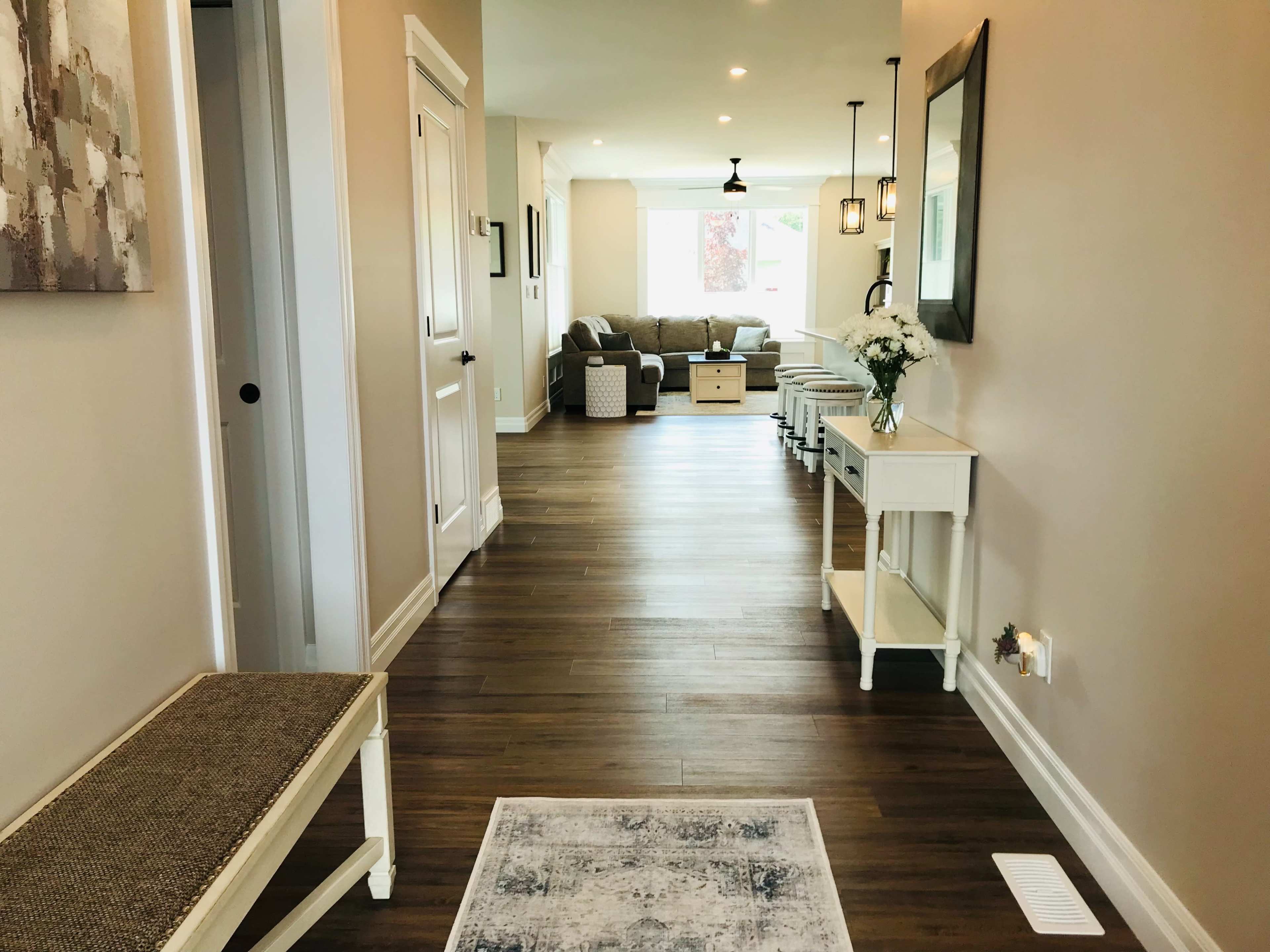 A hallway leads to a living area with a sofa, coffee table, and large windows, flanked by a bench and side tables.