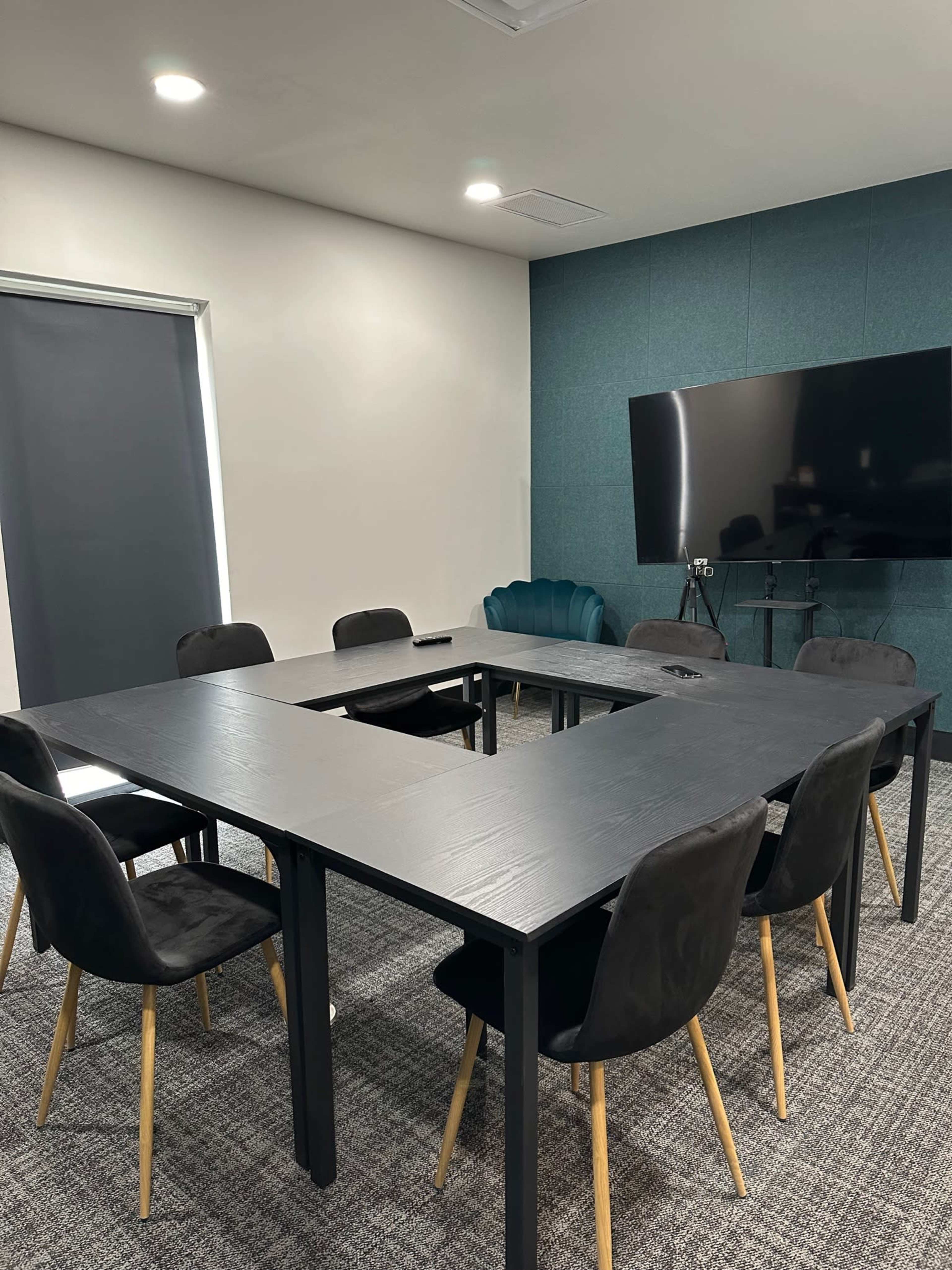 A modern conference room features a rectangular table surrounded by black chairs, with a large screen on one wall and a green accent wall.