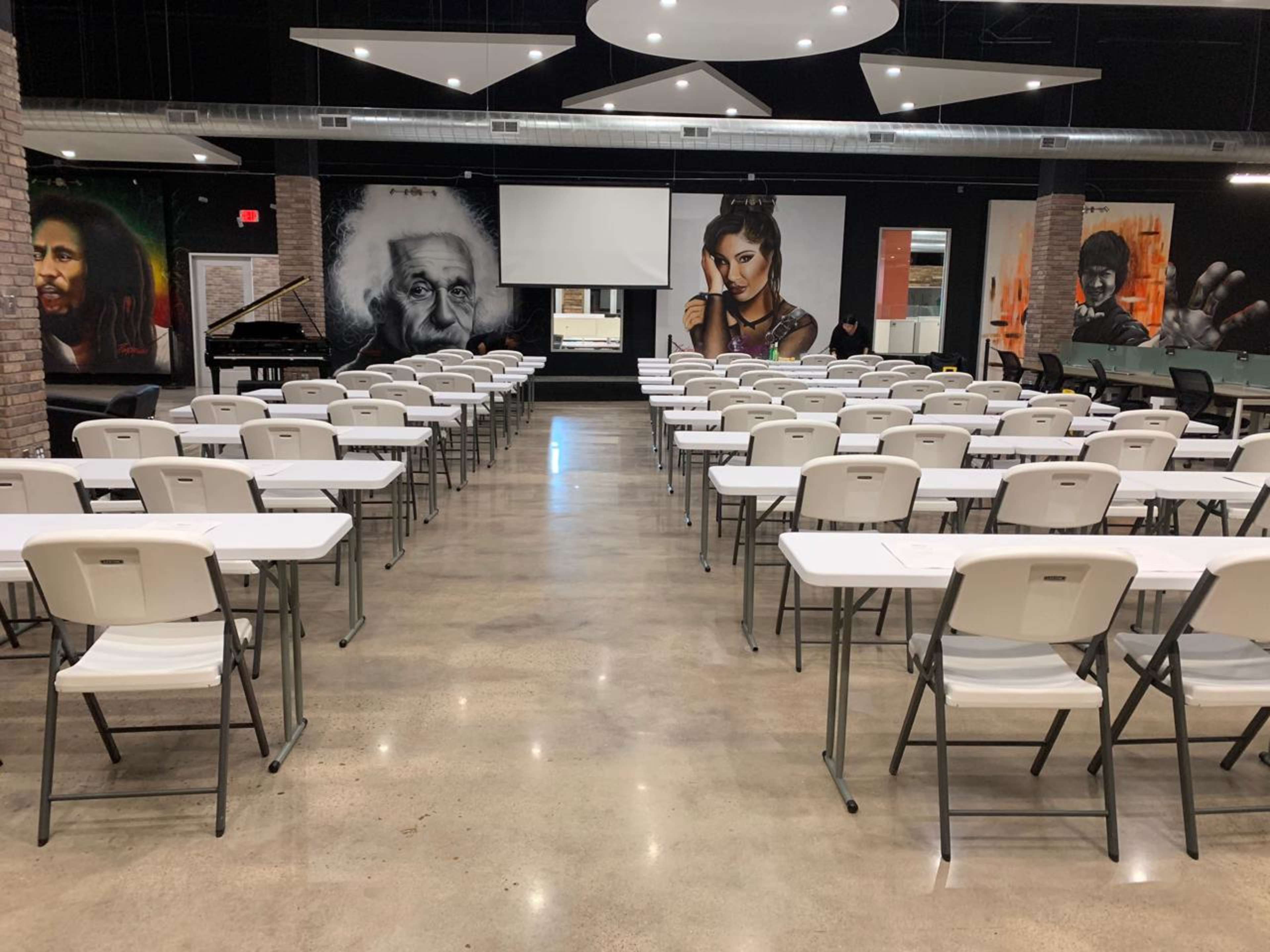 The image shows a spacious event room with rows of white folding tables and chairs, and large murals of notable figures on the walls.