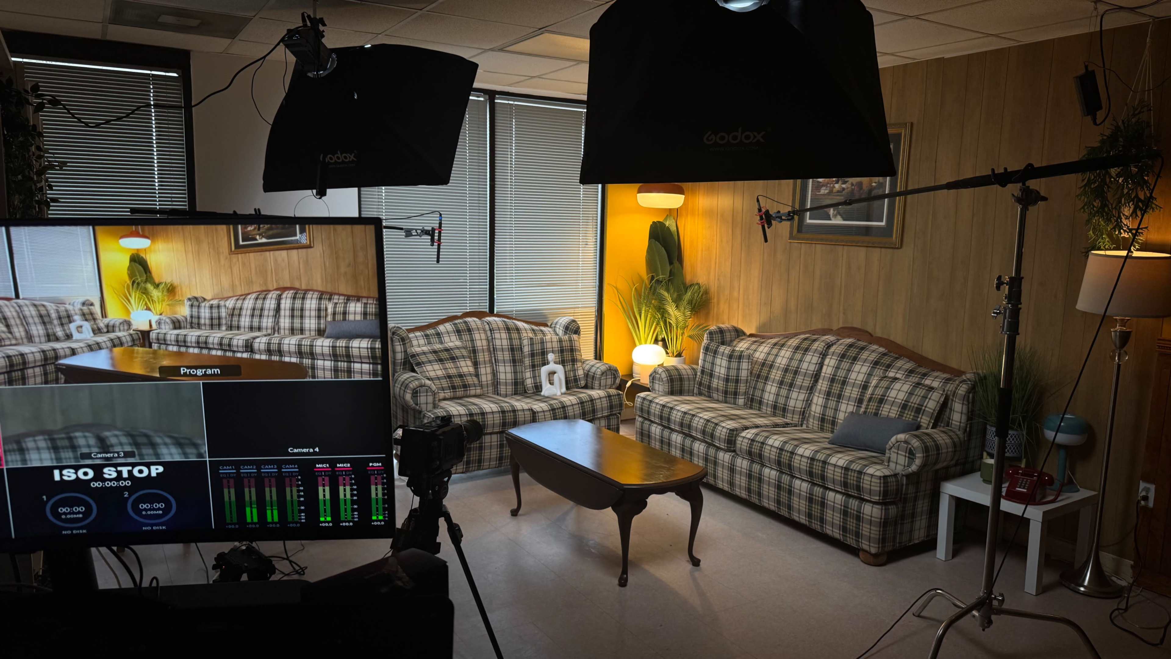 The image shows a well-lit studio setup featuring two plaid sofas, a wooden coffee table, and various lighting equipment in a room with large windows.