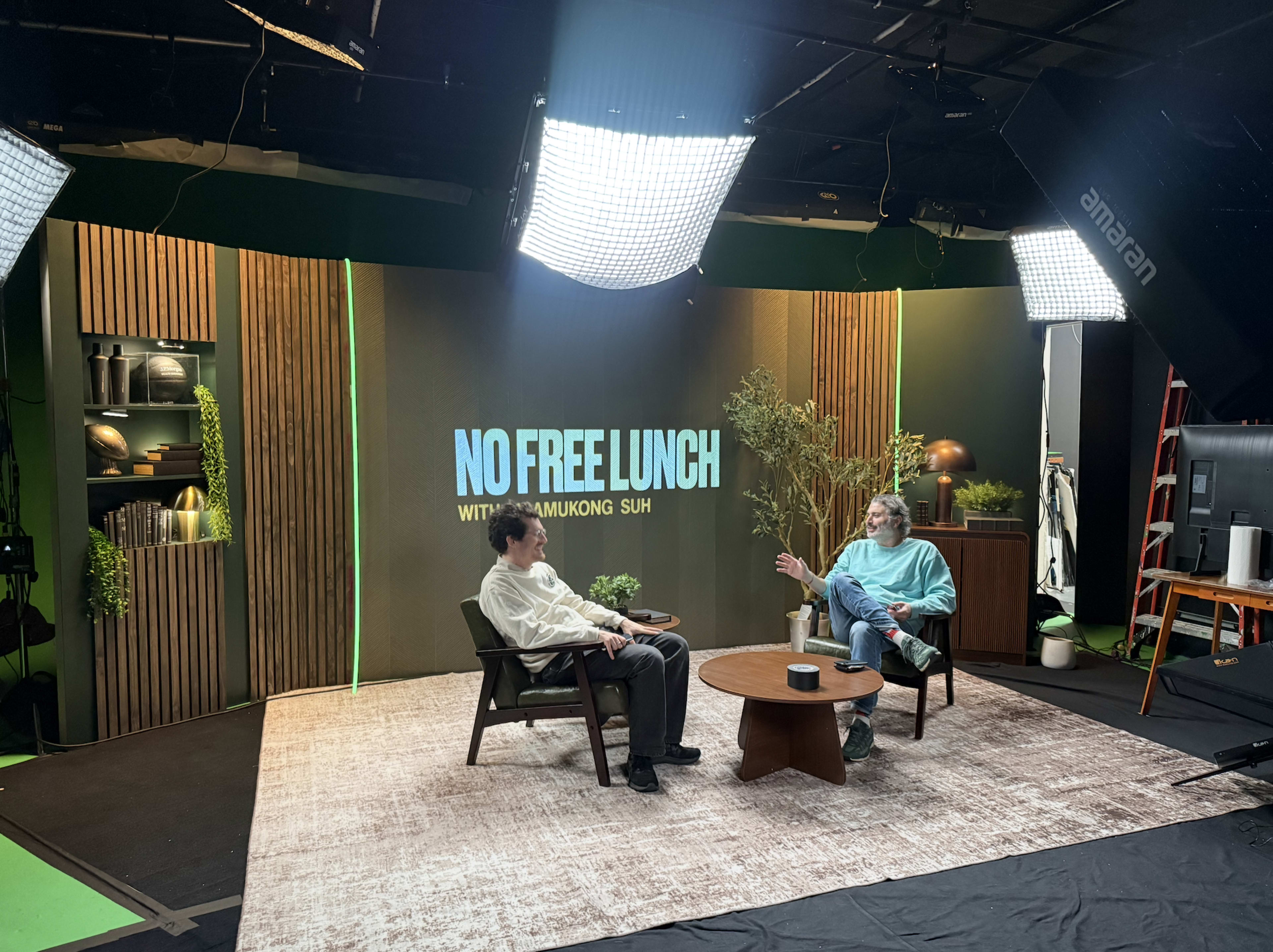 The image shows two men seated in an interview setting with a backdrop displaying the text "NO FREE LUNCH" and various decorative elements around them.