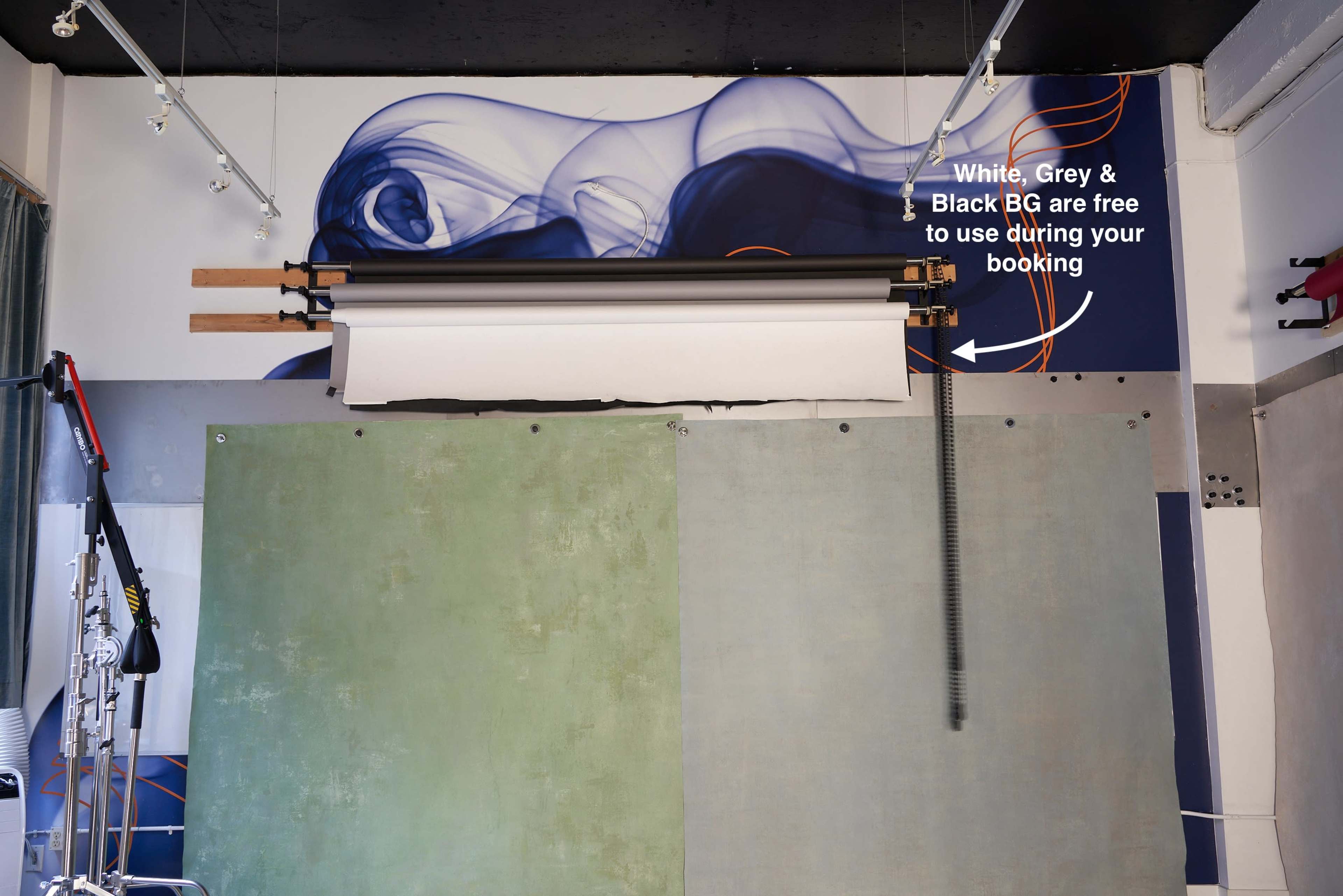 The image shows a photo studio setup featuring a roll-up backdrop system with white, gray, and black backgrounds mounted above textured green and blue walls.