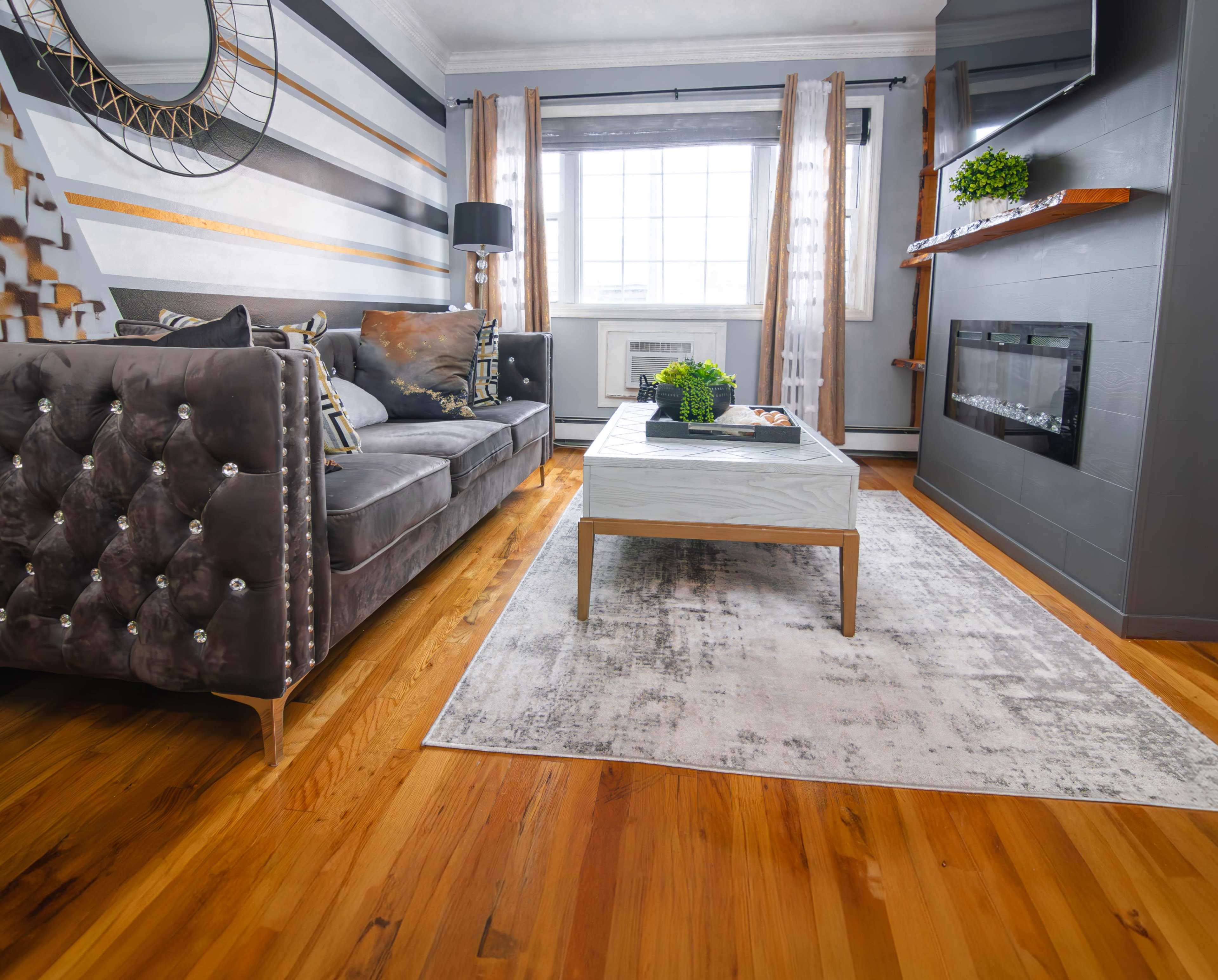 A modern living room features a gray tufted sofa, a white coffee table, and a wall-mounted television above a sleek fireplace, all set against hardwood floors and large windows.