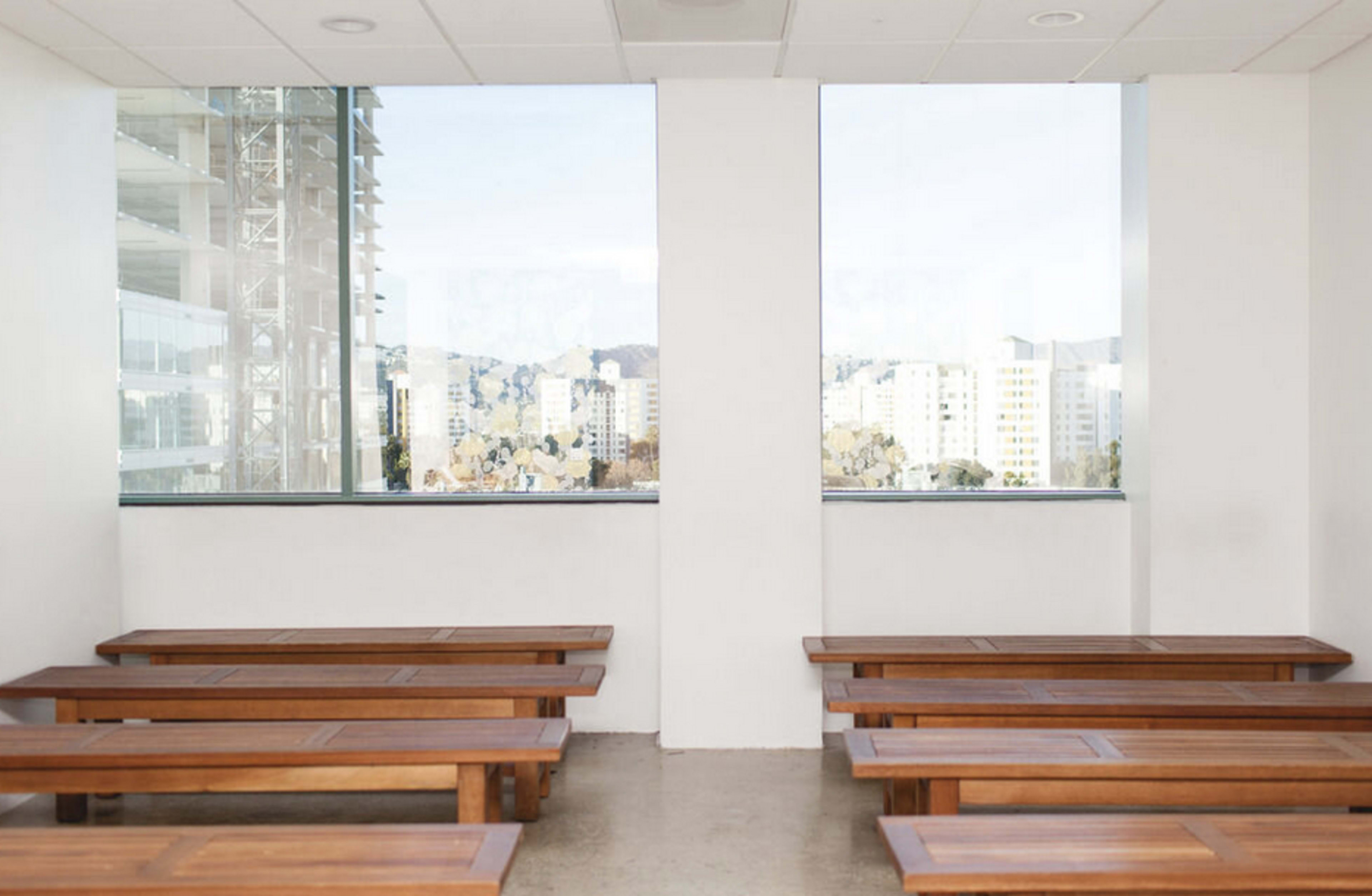 The scene shows a minimalist room with wooden benches arranged in two rows next to large windows overlooking a cityscape.
