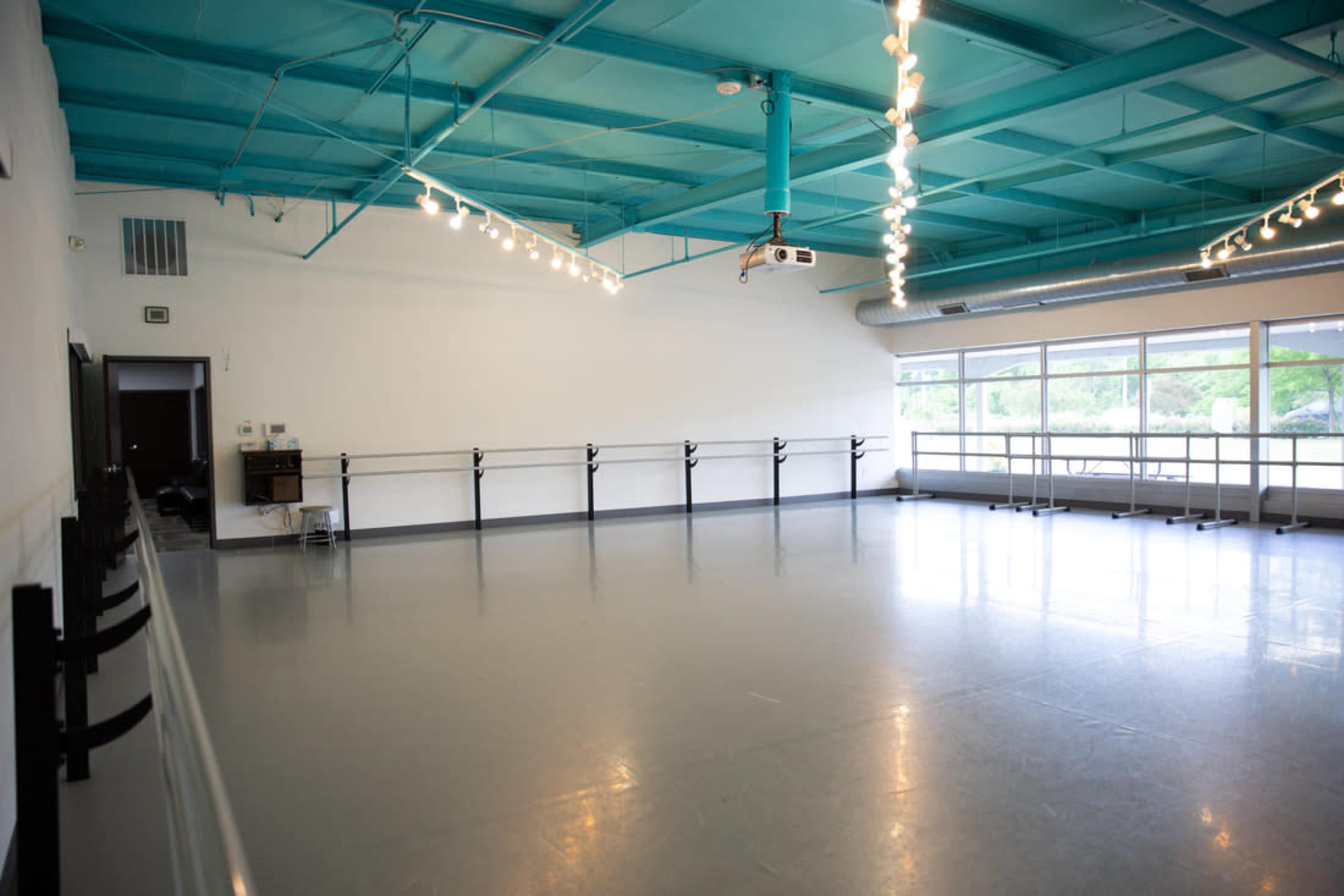 A spacious dance studio with a smooth gray floor, a turquoise ceiling, and large windows allowing natural light.