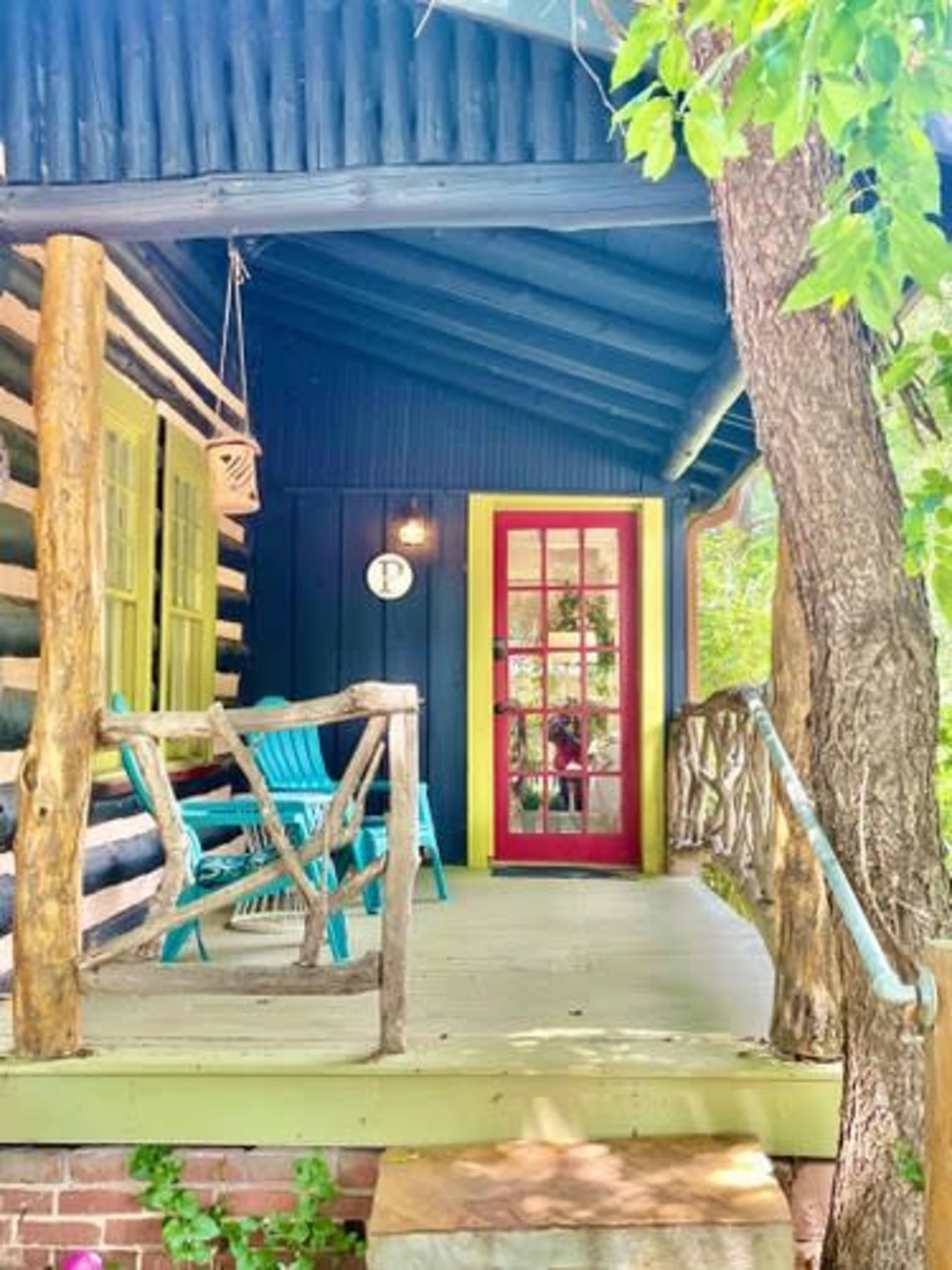 A charming porch with log walls, a red door, and blue chairs surrounded by greenery.