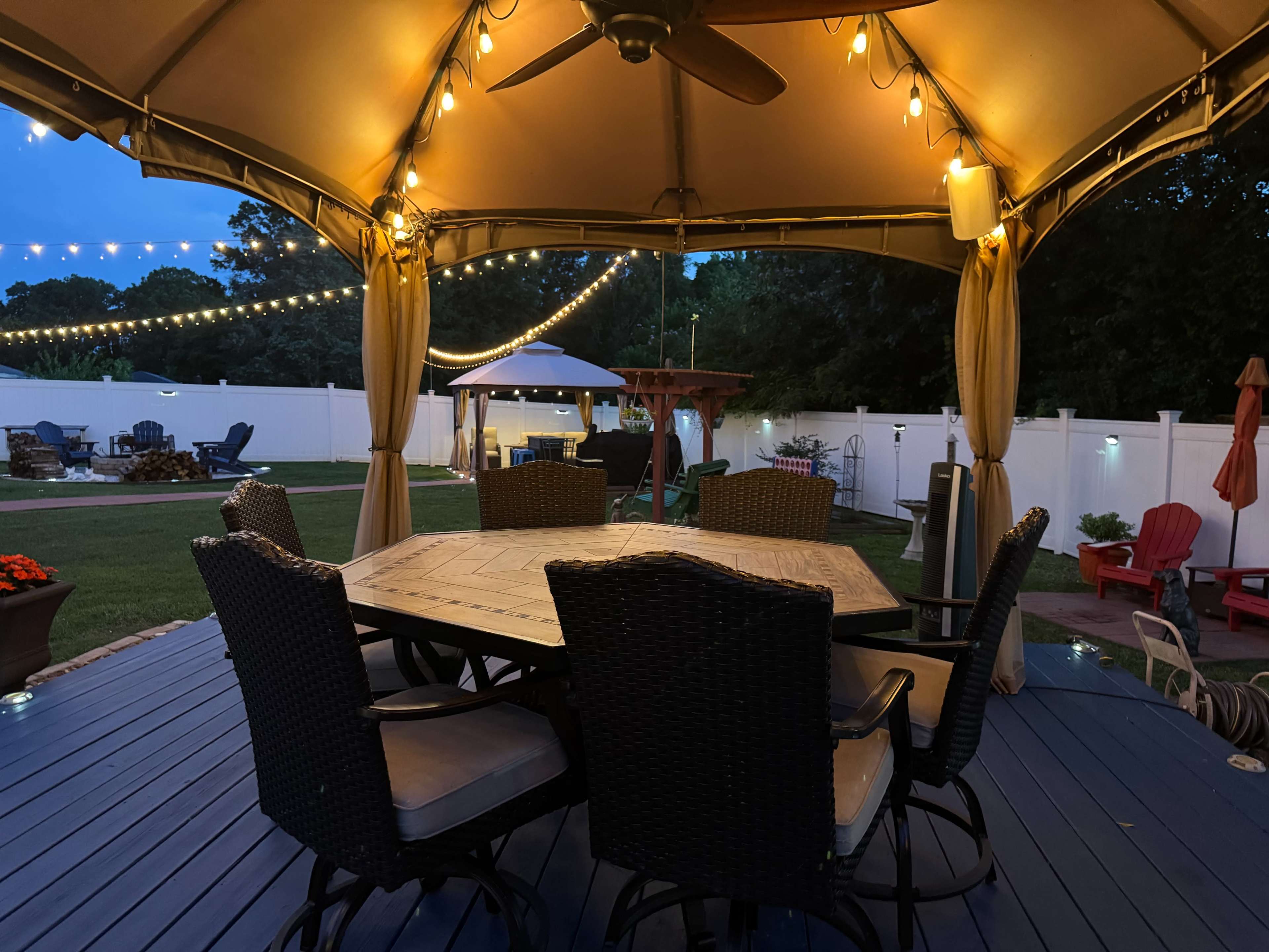 A covered outdoor seating area with a large table and chairs is illuminated by string lights, surrounded by a landscaped yard and additional seating arrangements.