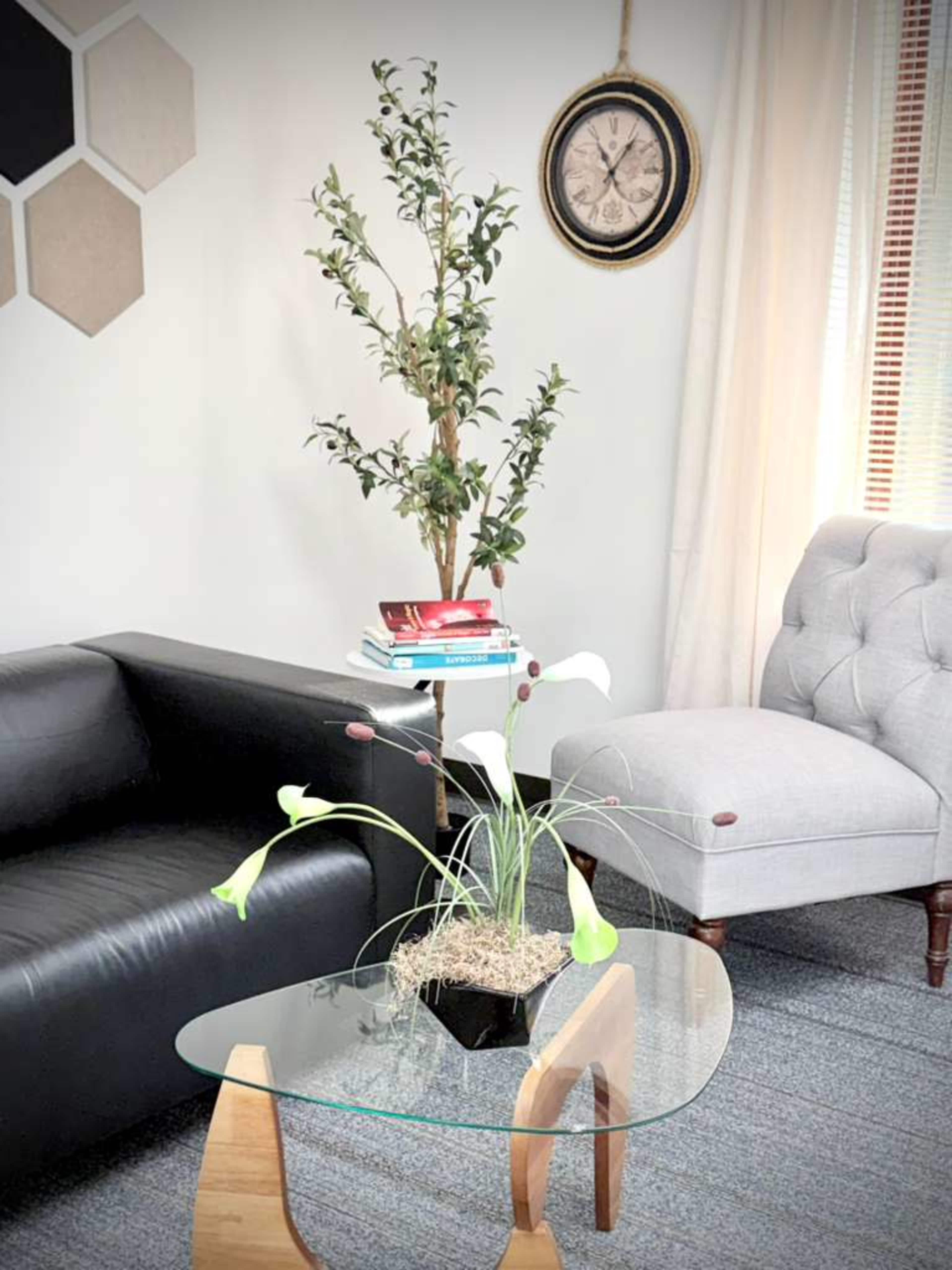 The image shows a modern living room setup featuring a black sofa, a gray armchair, a glass coffee table with a decorative plant, and geometric wall decor.