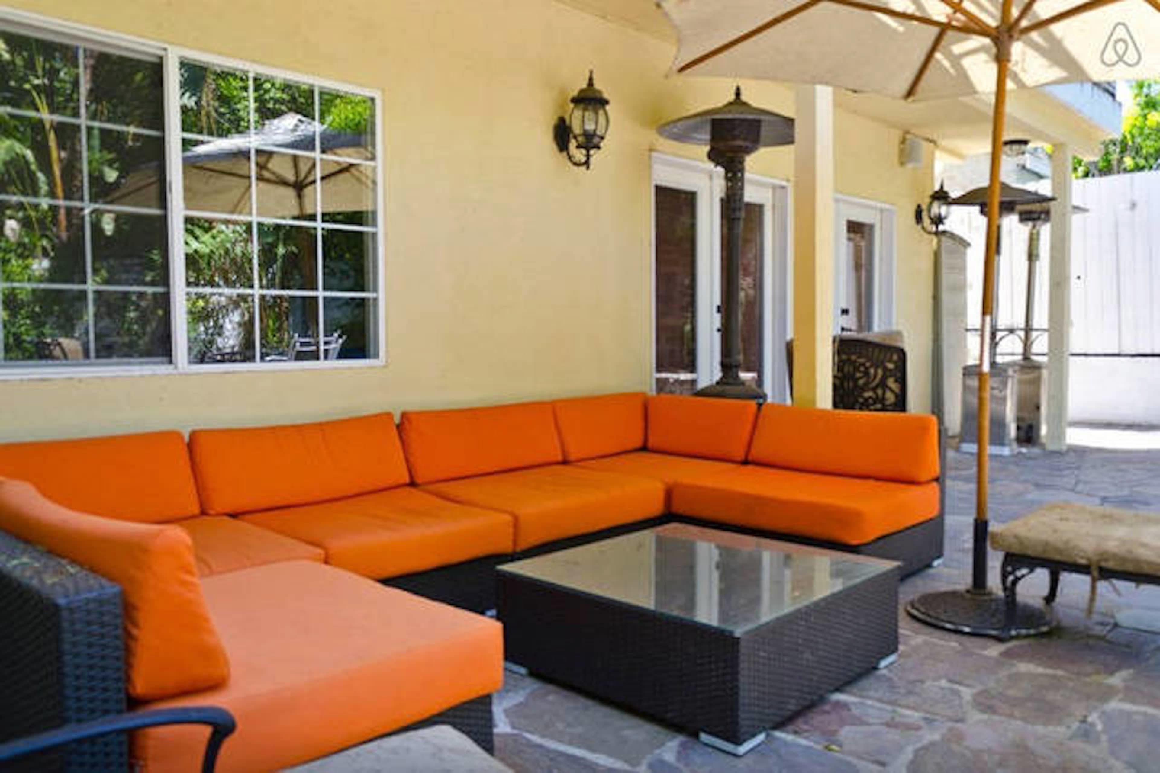 A large outdoor patio with a sectional orange sofa, a glass-top coffee table, and an umbrella providing shade.