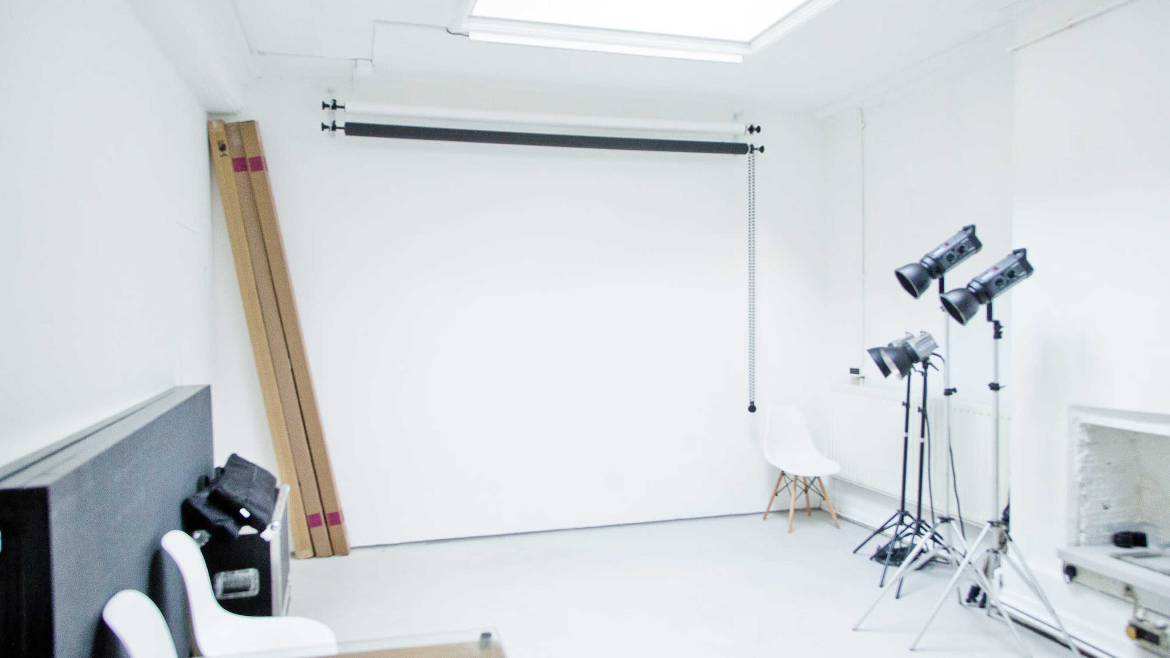 A minimalist photography studio with a white backdrop, lighting equipment, and a few chairs.