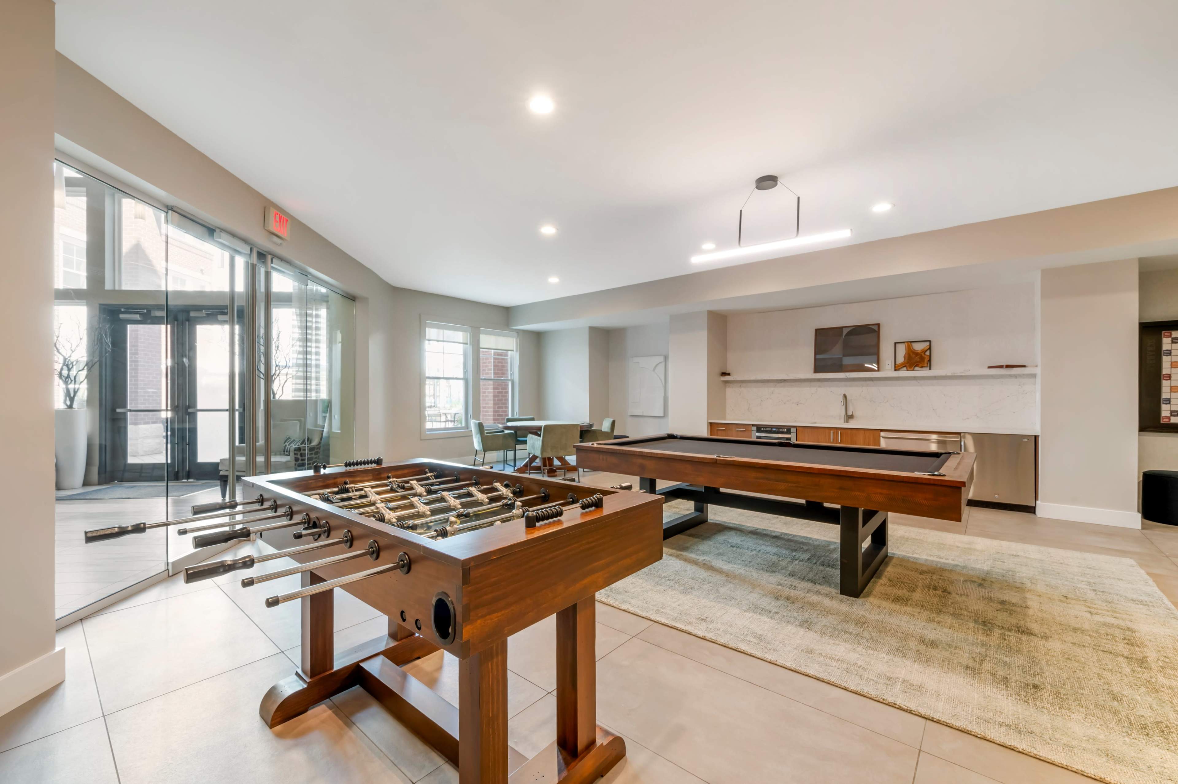 The image shows a recreational room featuring a foosball table, a pool table, and large windows allowing natural light to enter.