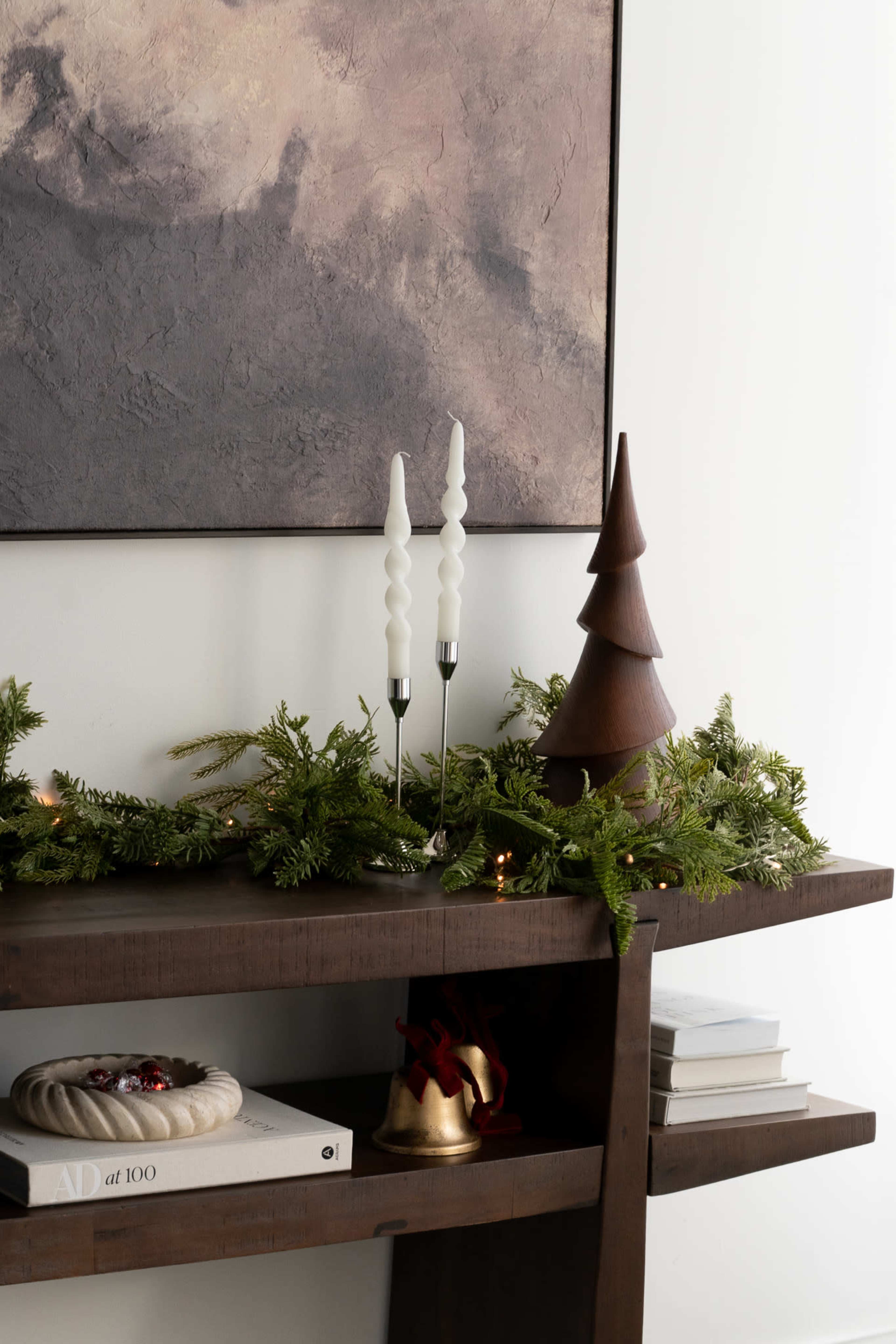A wooden shelf displays a holiday arrangement with green garlands, two white candlesticks, a wooden tree sculpture, and several books.