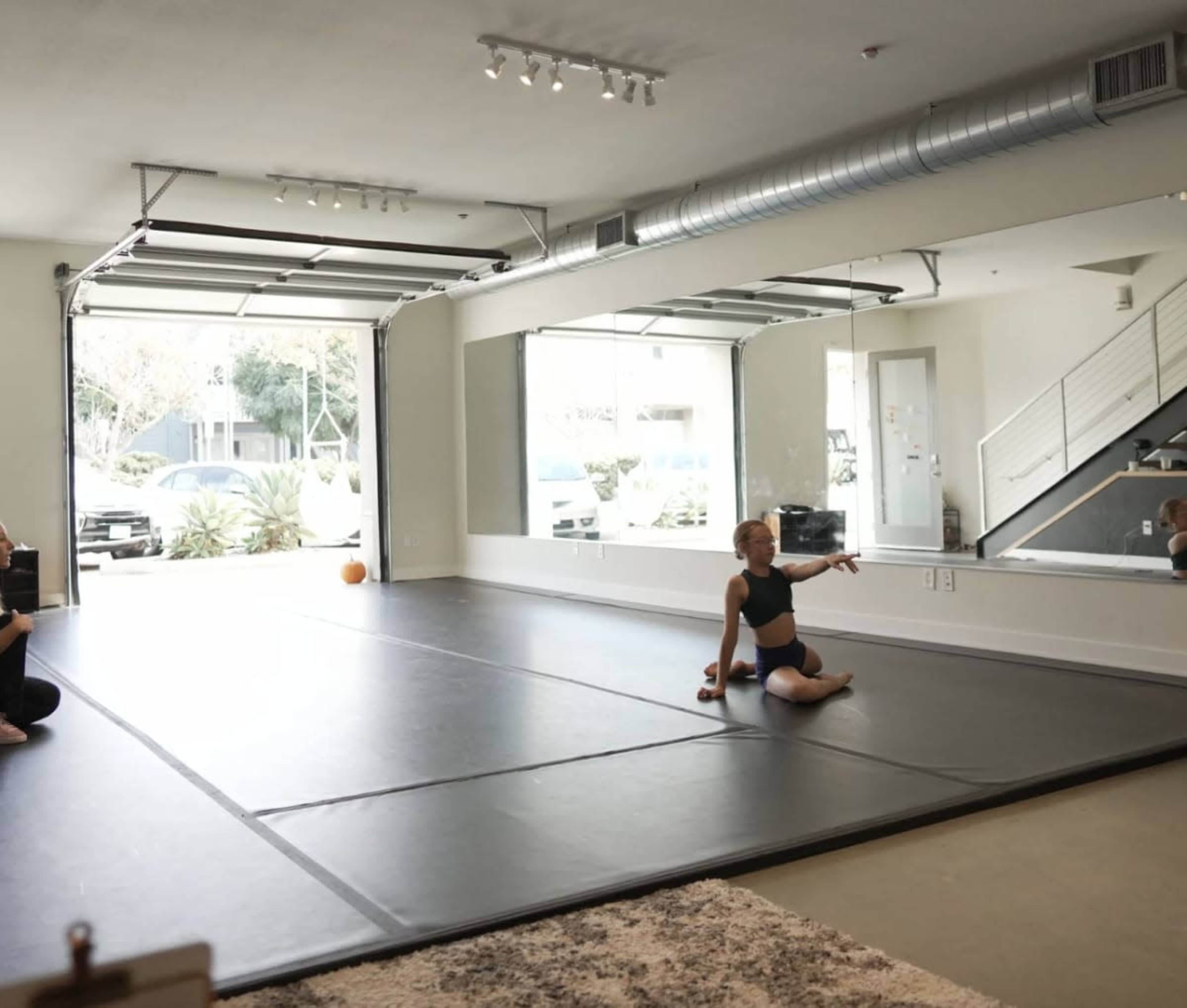 A dancer performs on a padded floor in a spacious studio featuring large mirrors and a garage door, with a person observing from the side.