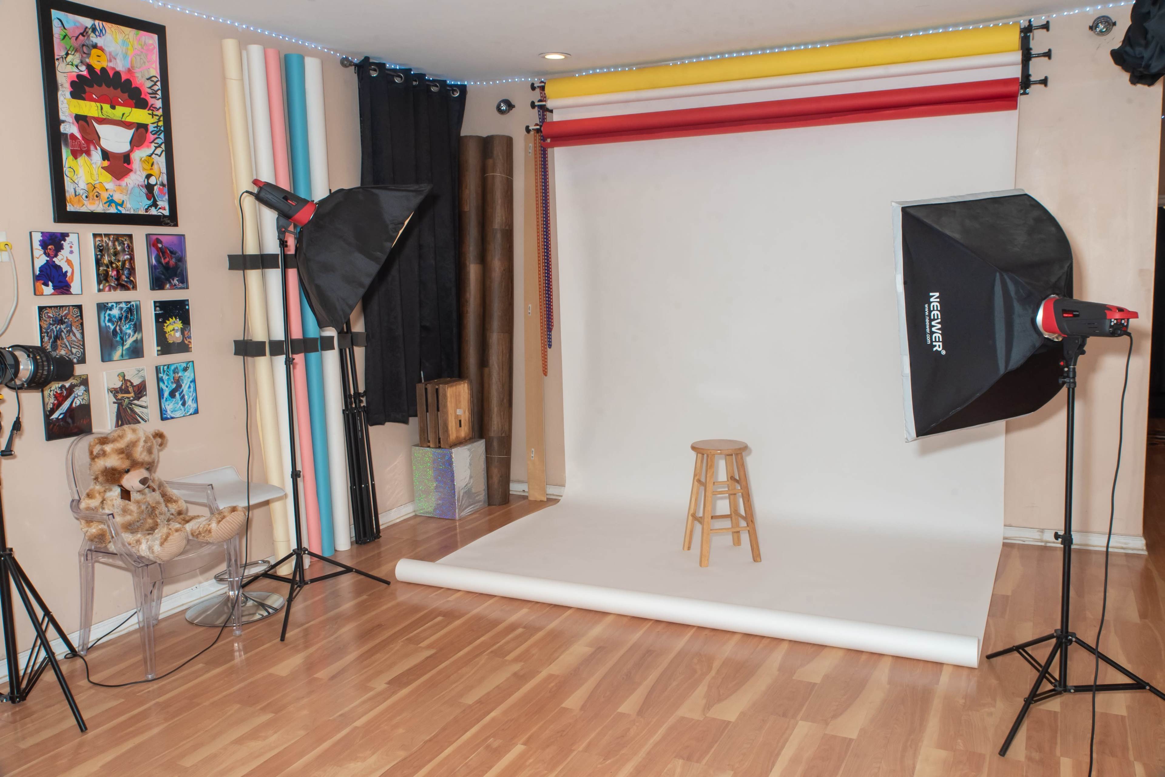 The image shows a photography studio with a white backdrop, a wooden stool, two studio lights, and colorful backdrops on the wall.